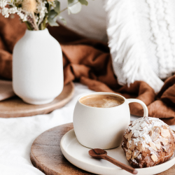A white mug of coffee, a pastry dusted with powdered sugar, and a spoon on a wooden tray, with a vase of flowers in the background.