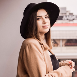 A woman wearing a black wide-brimmed hat and tan coat, looking at the camera with a slight smile, standing indoors.