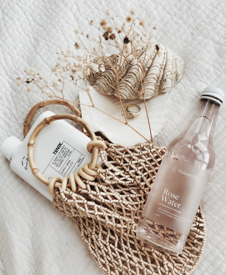 A woven handbag containing a roll of toilet paper, a white bottle, and dried flowers. Next to the bag is a bottle of Madame Brew Rose Water on a white textured surface with a seashell and a ring nearby.