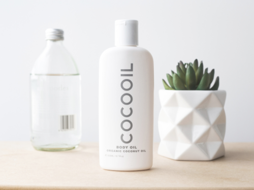 A bottle of body oil labeled 'COCOOIL' placed on a wooden surface next to a small plastic water bottle and a pot with a green succulent plant.