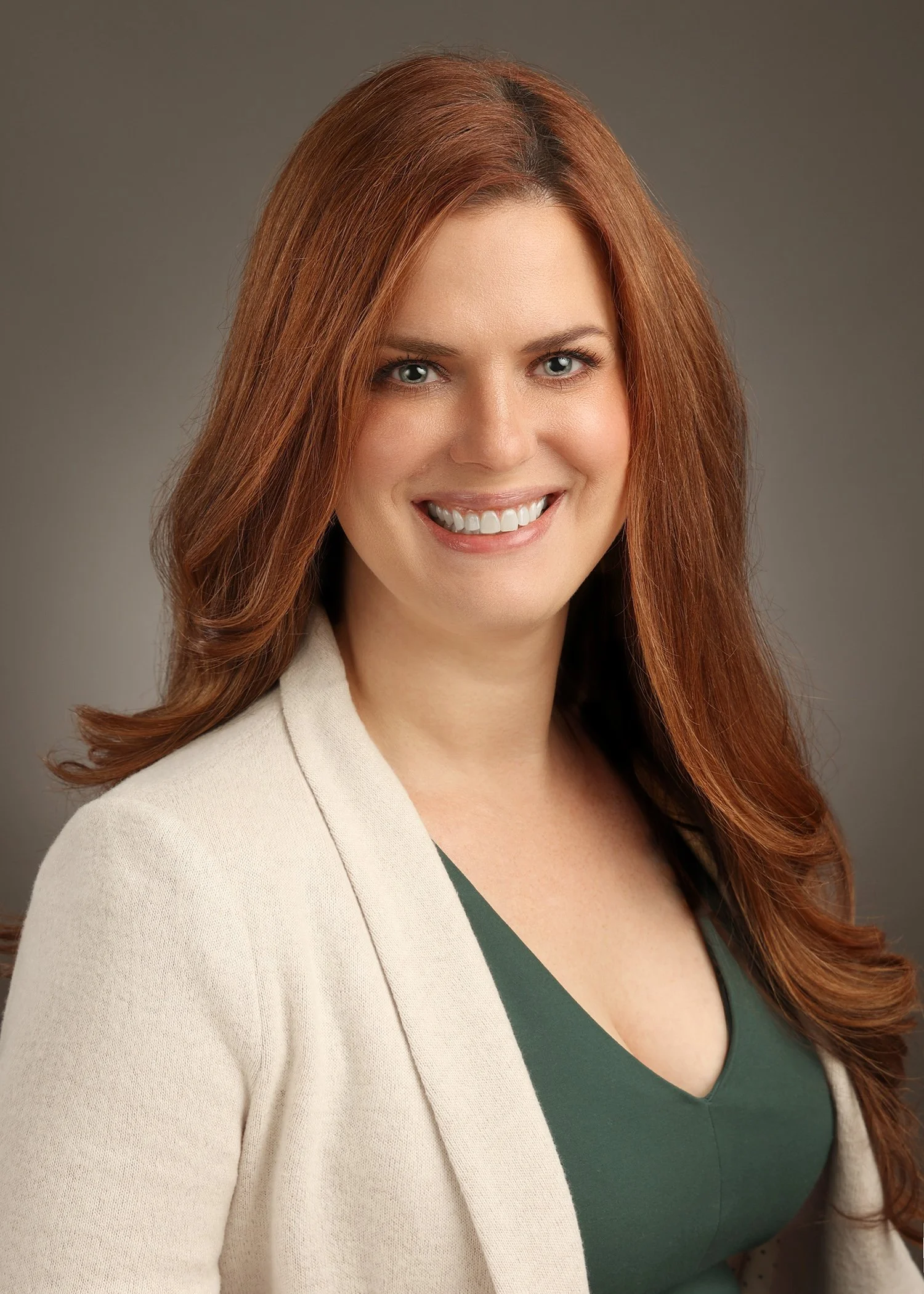A woman with long, wavy auburn hair smiling and wearing a beige blazer over a dark green top.