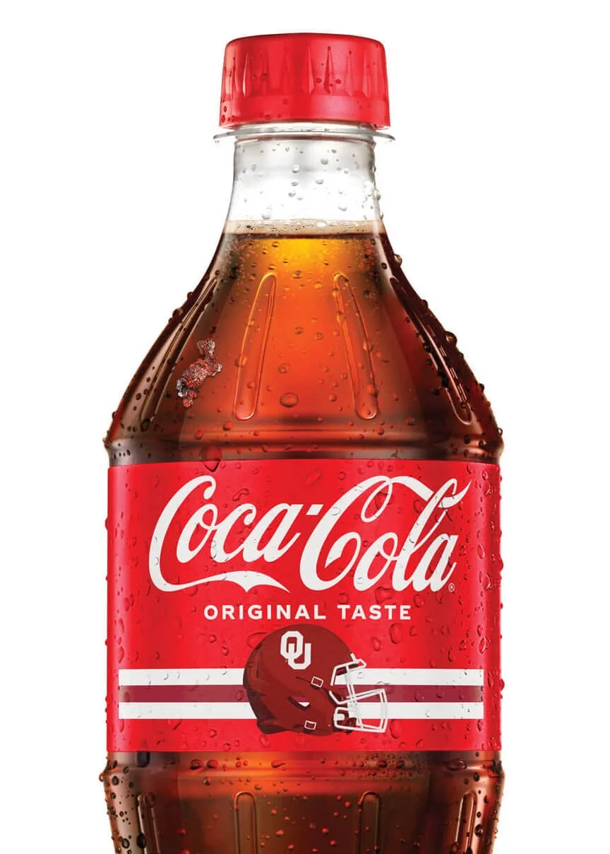 A plastic bottle of Coca-Cola with condensation droplets, featuring a red label with the Coca-Cola logo, and an Oklahoma Sooners football helmet graphic on the label.