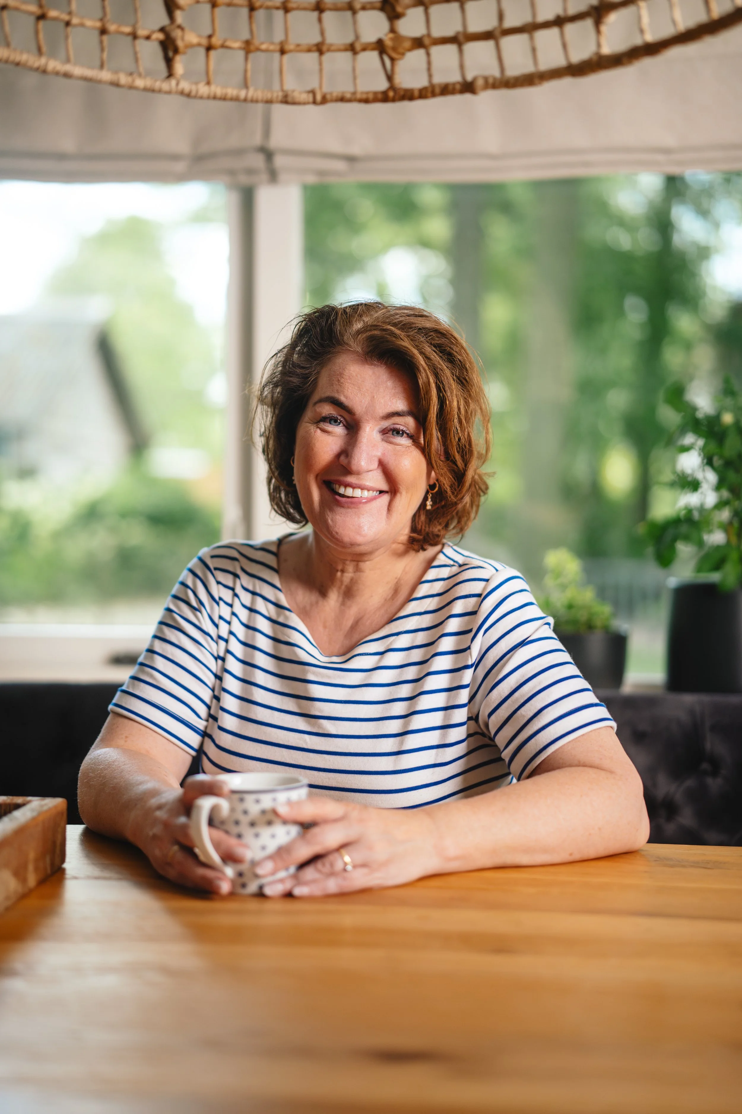 Een vrouw met bruin haar en een blauwe en witte gestreepte shirt die in een keuken zit, lachend met een mok in haar handen, met een raam en groene tuin op de achtergrond.