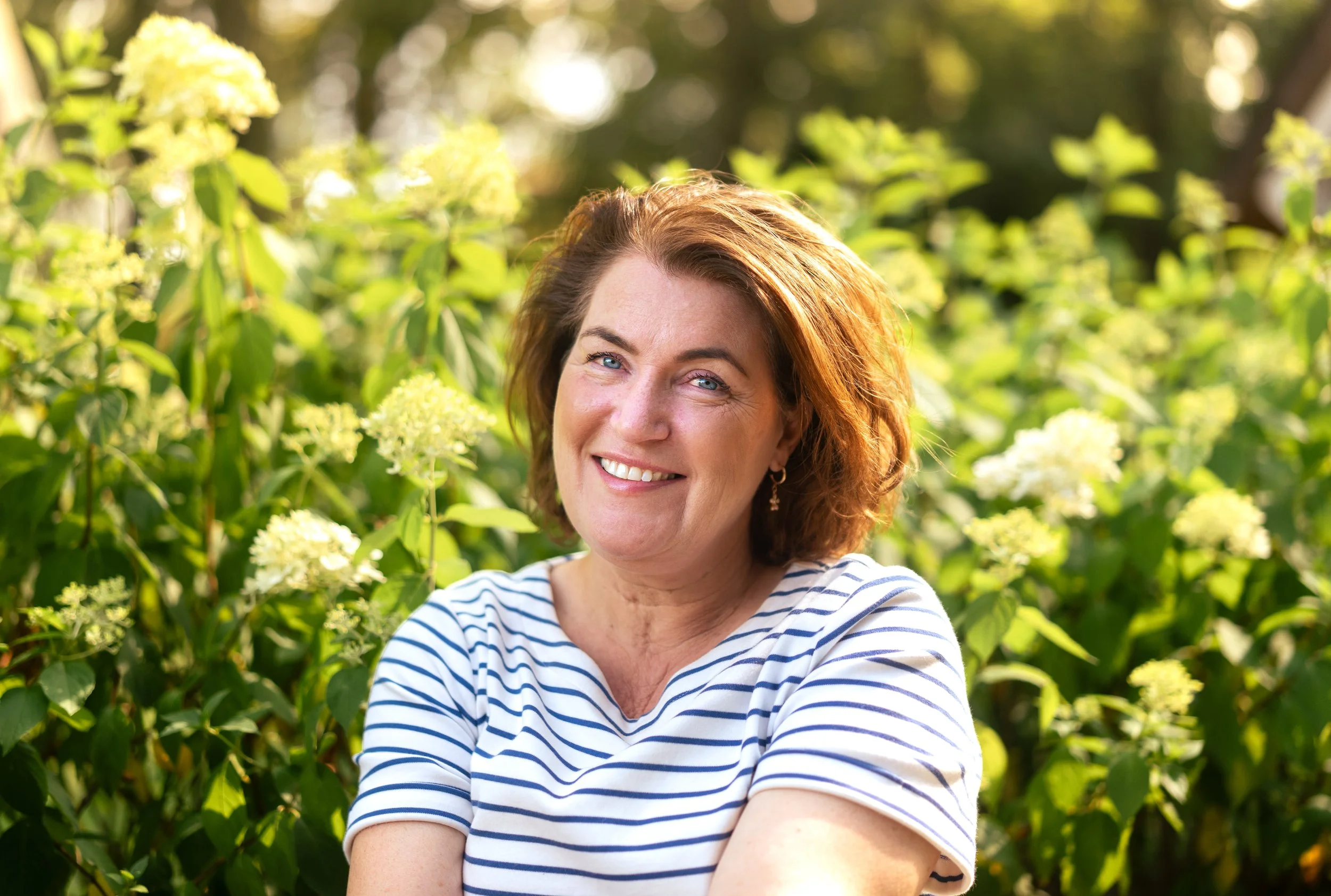 Vrouw met bruin haar en een witte en blauwe gestreepte top die lachend poseert in een tuin met groene planten en bloemen.