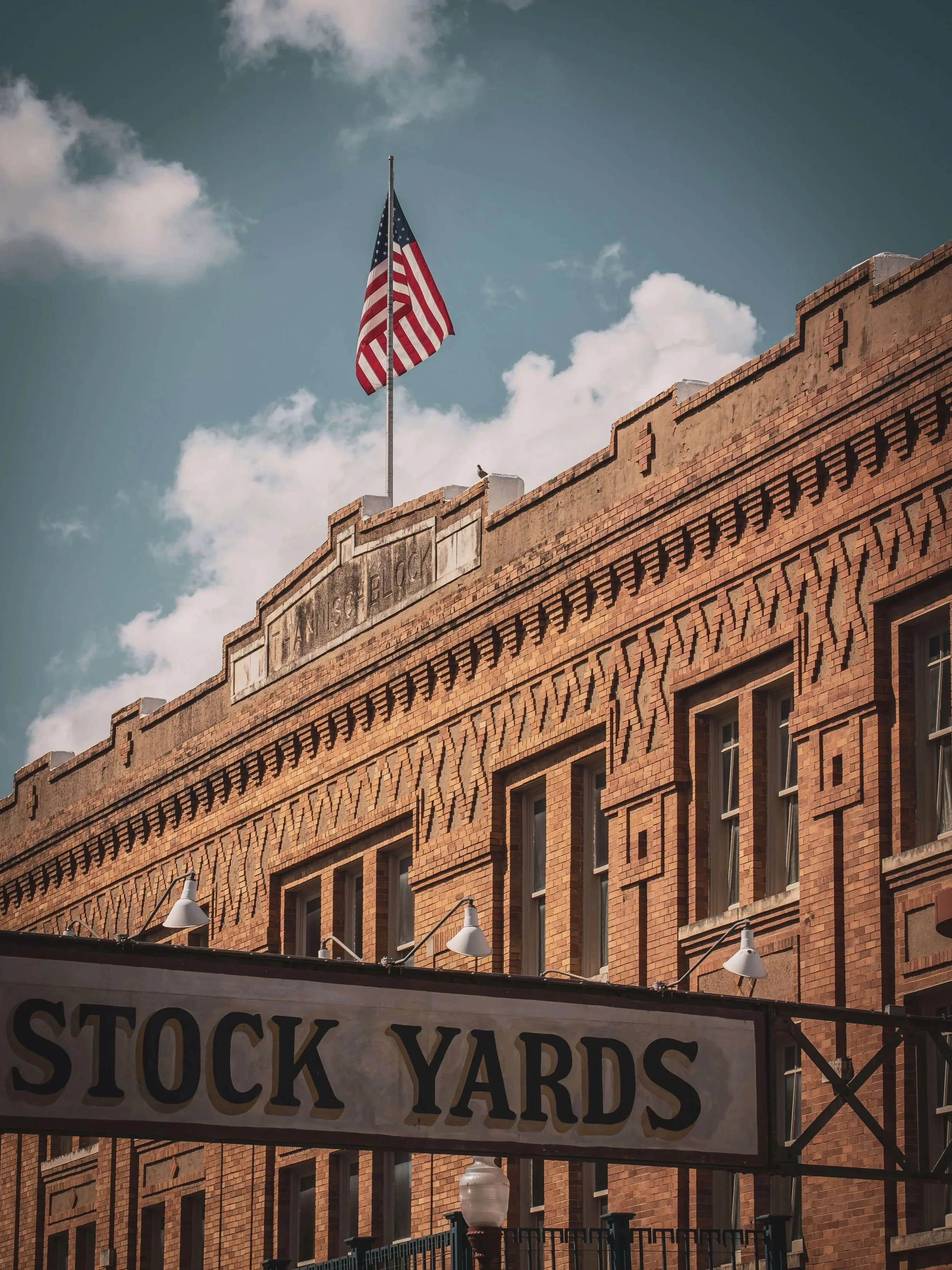 Historic Fort Worth Stockyards brick building sign flying an American flag under a cloudy blue sky.