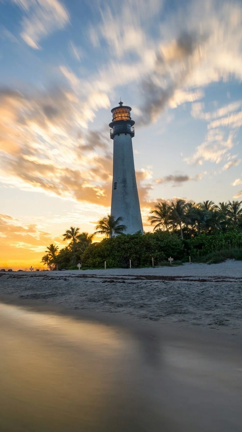 A lighthouse standing tall on a beach with palm trees, during sunset with colorful clouds in the sky.