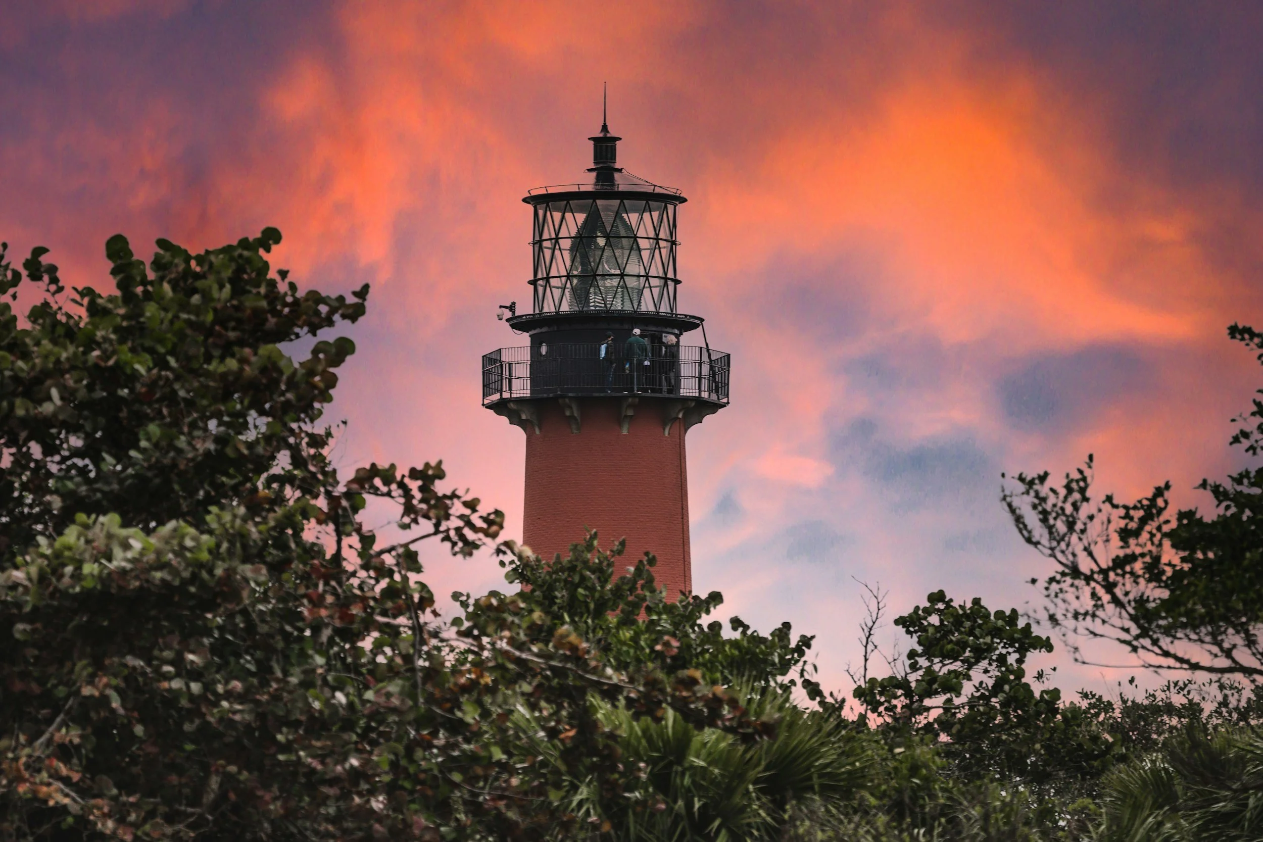 A lighthouse tower with a balcony and glass windows at the top, set against a colorful sunset sky with pink and orange clouds, surrounded by green leafy trees.