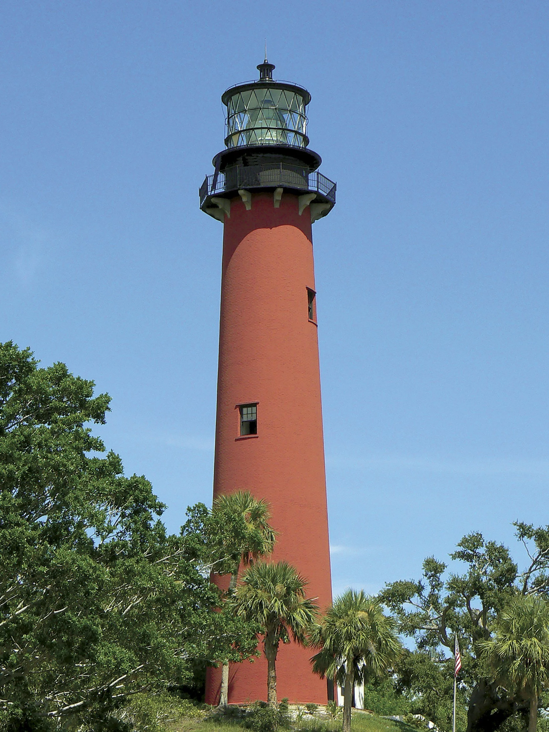 A red lighthouse with a black and clear glass top, surrounded by trees and greenery against a blue sky.