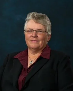 A professional portrait of an older woman with short gray hair, glasses, wearing a dark blazer over a maroon blouse, against a dark blue background.