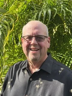 A smiling man with glasses and a bald head standing outdoors among green tropical plants.