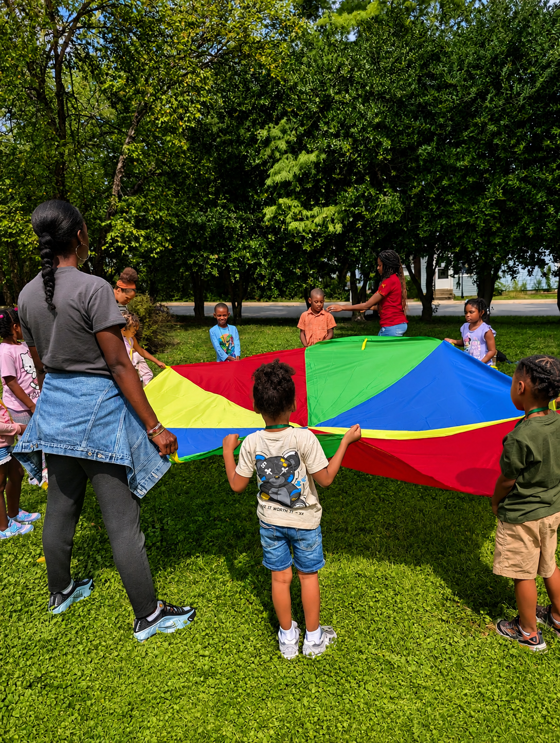 Children and adults playing with a colorful parachute outdoors on green grass with trees in the background.