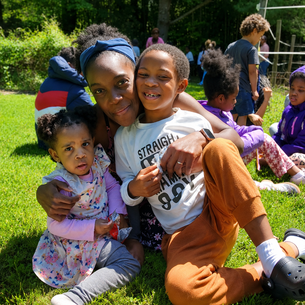 A woman with two young children sitting on grass at an outdoor gathering, smiling and hugging. Other children are in the background, sitting and interacting.