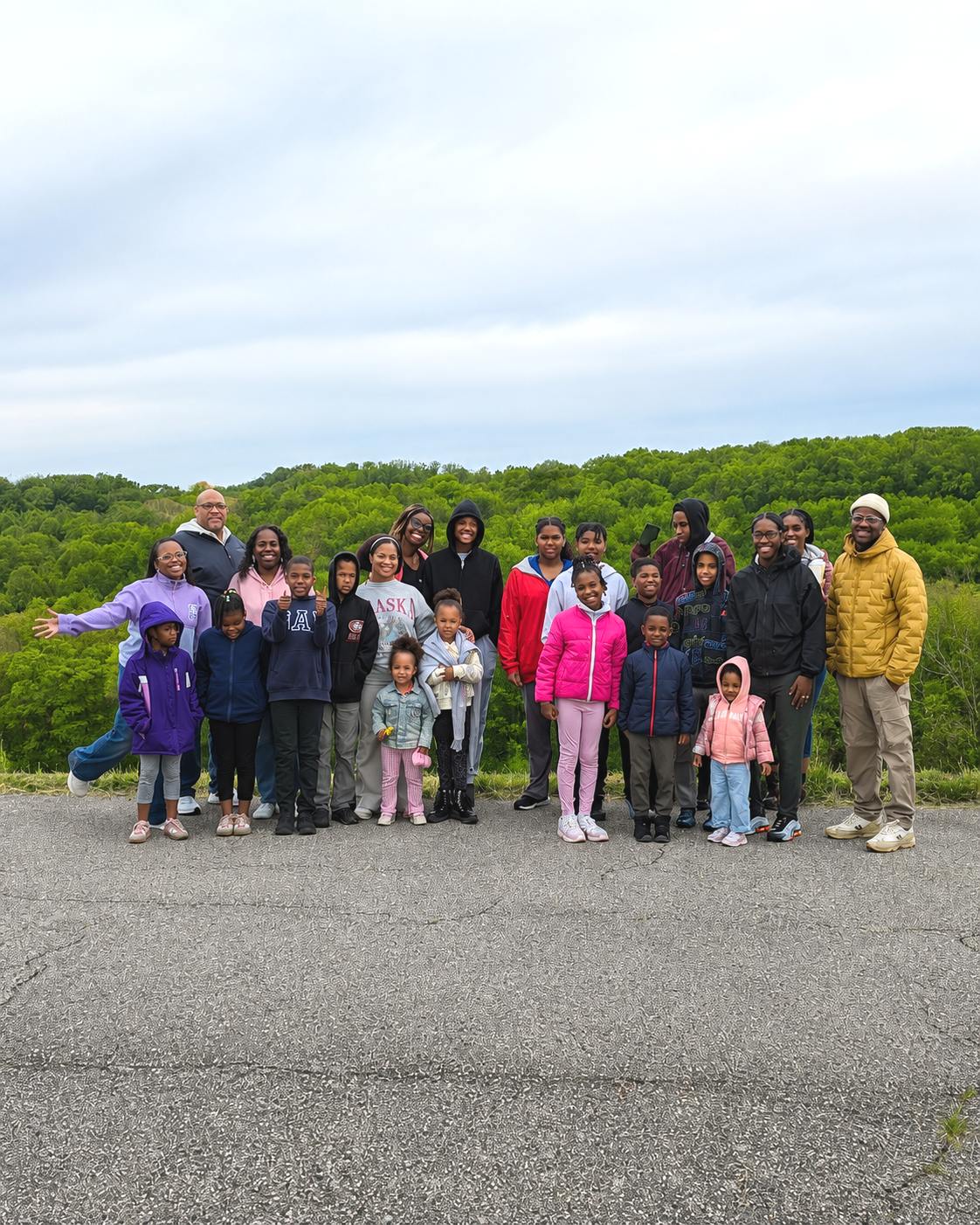 Group of diverse children and adults outdoors on a cloudy day with green hills in the background.