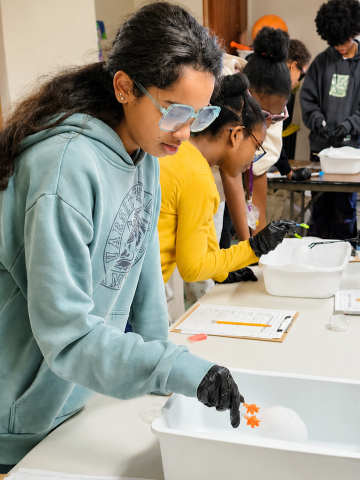 Young girl with glasses and black gloves participating in a science activity, handling a foam and small orange toy in a white container, with other students working at tables in the background.