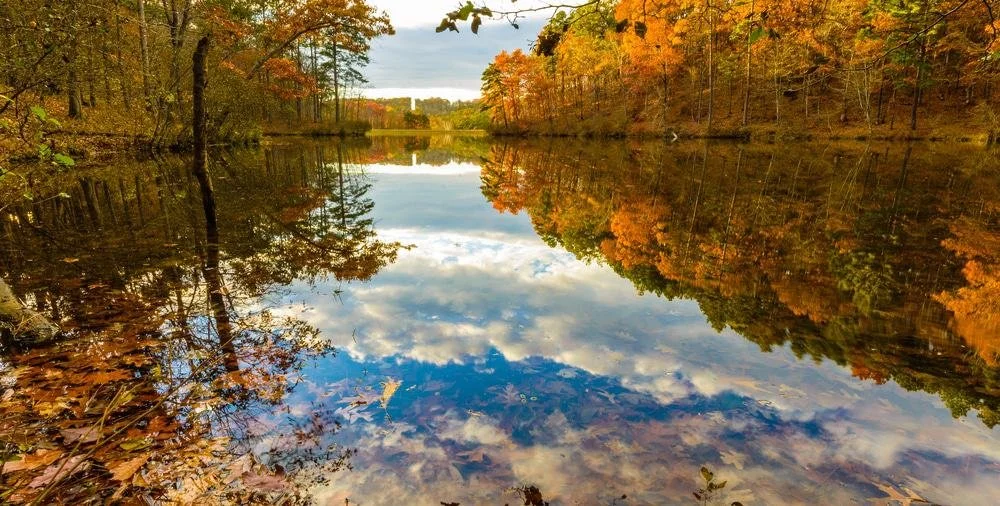 A peaceful autumn scene with trees with colorful fall foliage reflecting on a calm river under a partly cloudy sky.