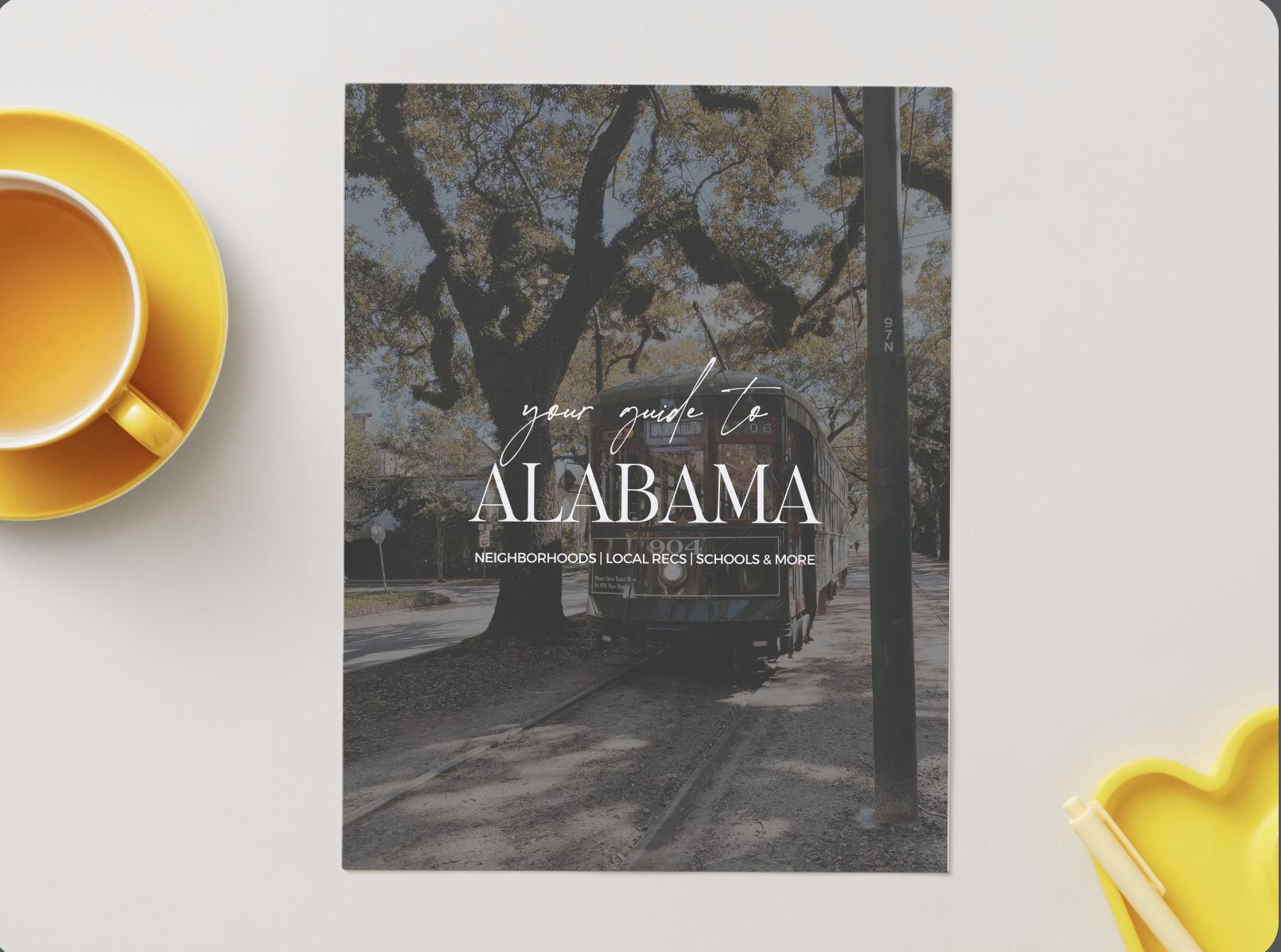 A travel guidebook titled "Your Guide to Alabama" with a vintage streetcar on the cover, surrounded by a yellow cup of tea on a white surface.