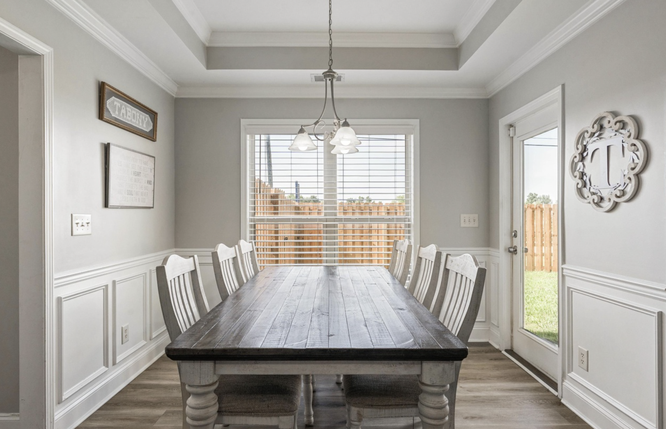 A dining room with a large wooden table surrounded by white chairs, white paneled walls, large windows with shutters, and a view of a backyard with a wooden fence.