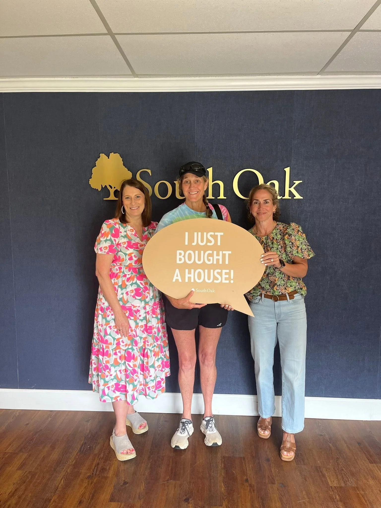Three women standing in front of a dark blue wall with a gold South Oak logo. One woman is holding a sign that says 'I JUST BOUGHT A HOUSE!'. All are smiling and dressed in casual summer attire, wearing sandals and athletic shoes.