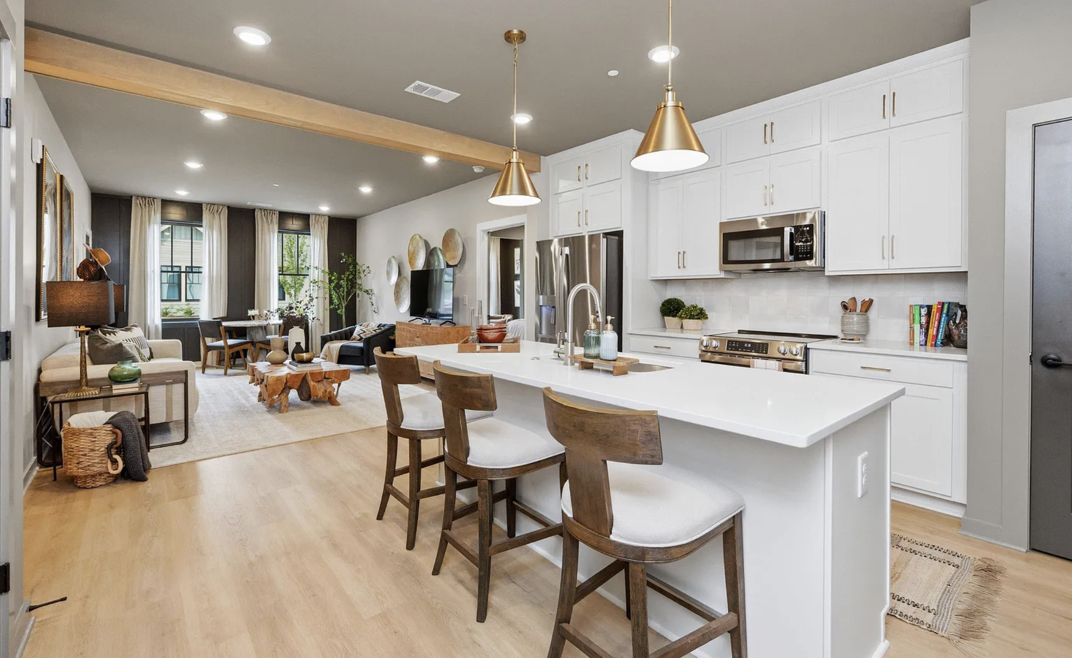 Open-concept living room and kitchen with white cabinetry, stainless steel appliances, pendant lights, wooden bar stools, and furniture including a sofa, armchairs, and coffee table, with large windows and decorative wall art.