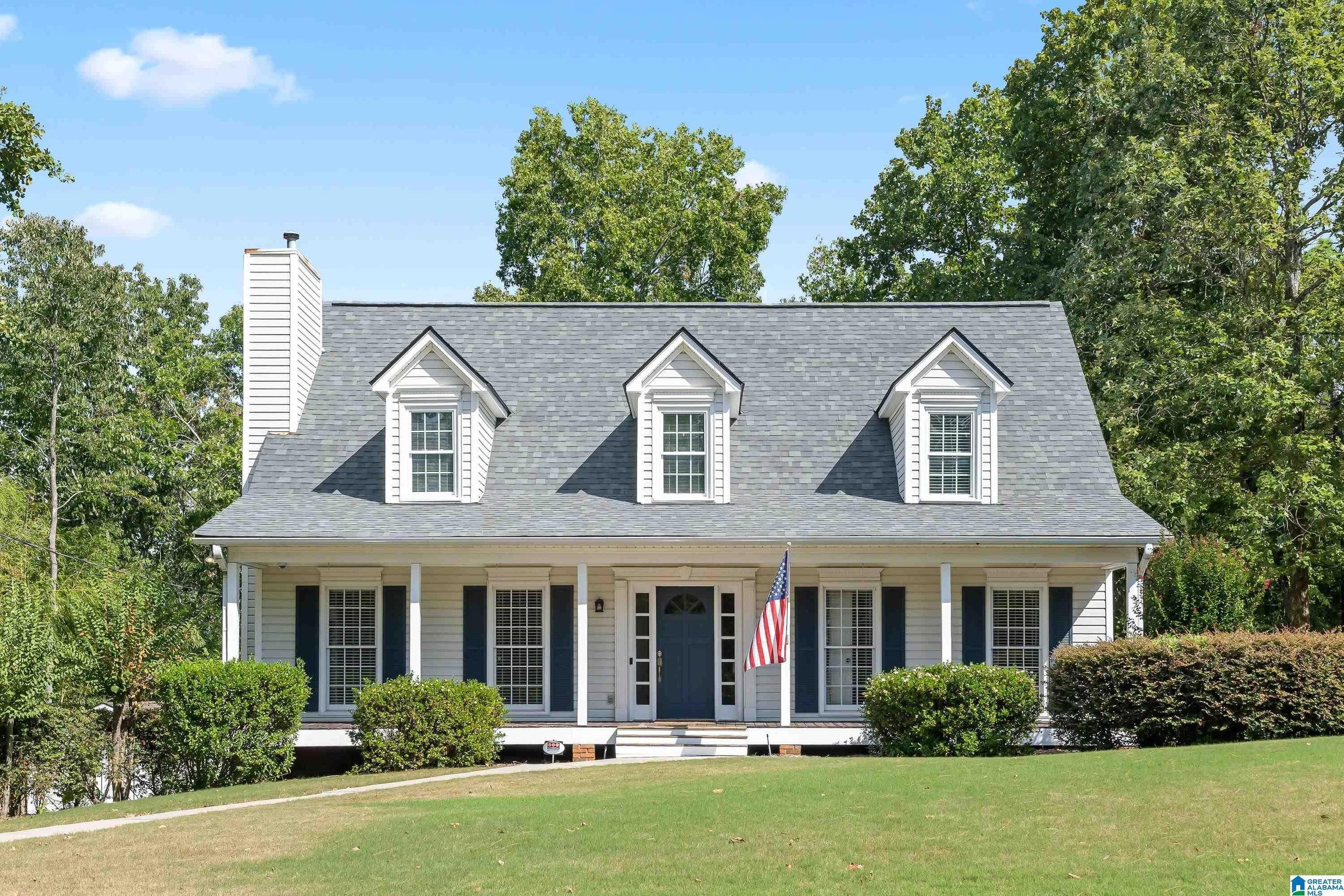 A large white house with a gray shingle roof, three dormer windows, and a front porch with an American flag, surrounded by green trees and a well-maintained lawn.