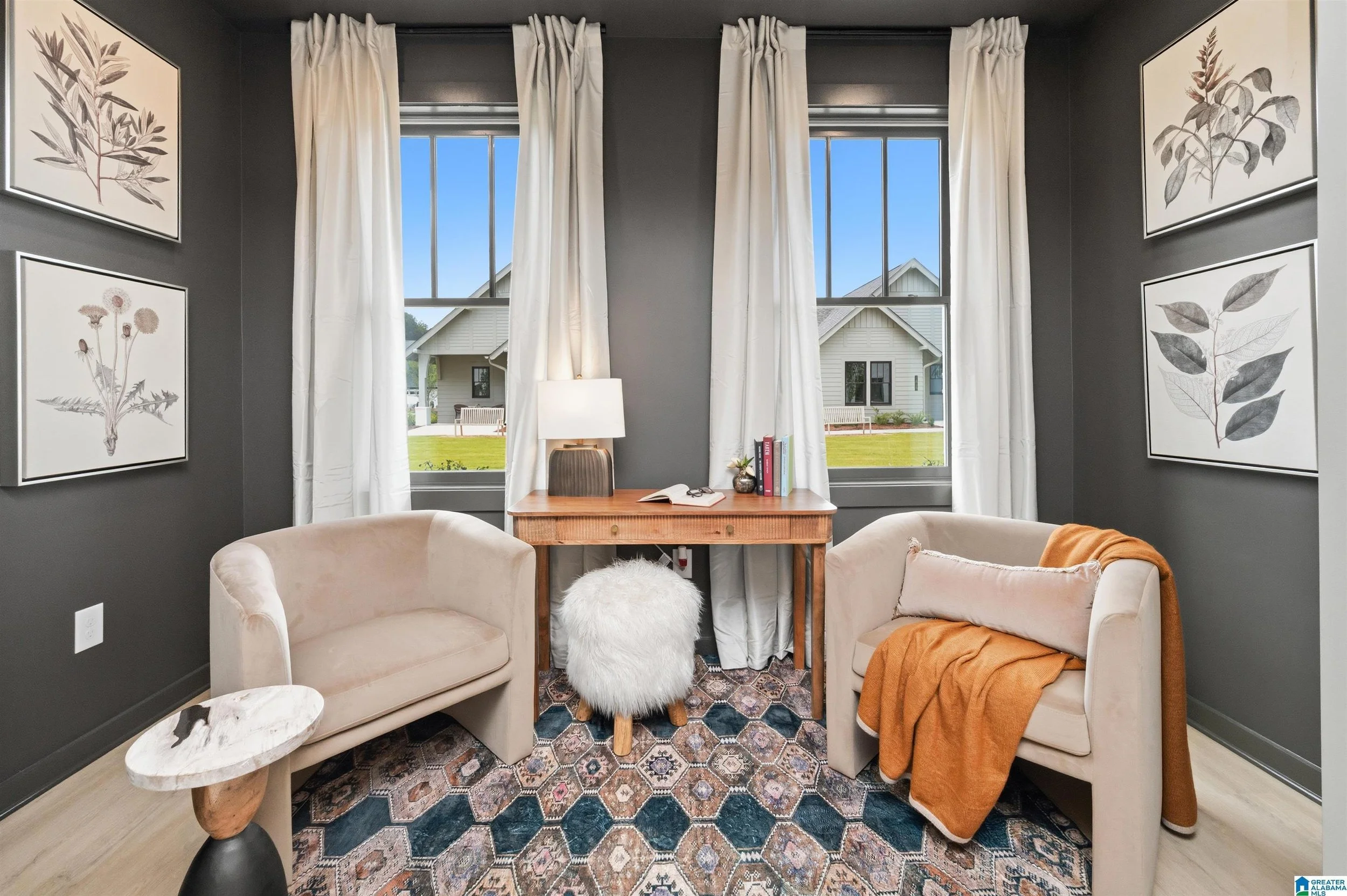Living room with dark gray walls, large windows with white curtains, cream-colored armchairs, a wooden table in between, a white fluffy stool, artwork of botanical illustrations on the walls, and a patterned rug.