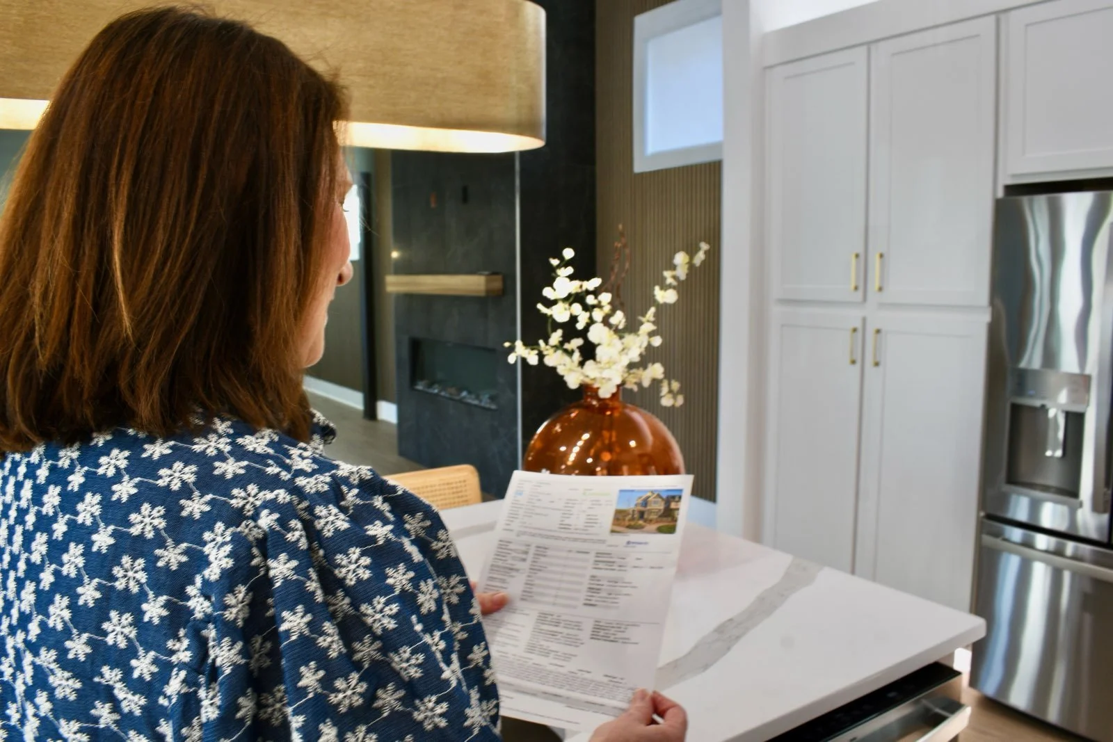 Woman with brown hair in a blue dress with white floral pattern holding a paper, standing in a modern kitchen with white cabinets, stainless steel refrigerator, and a large brown vase with white flowers on a white marble countertop.