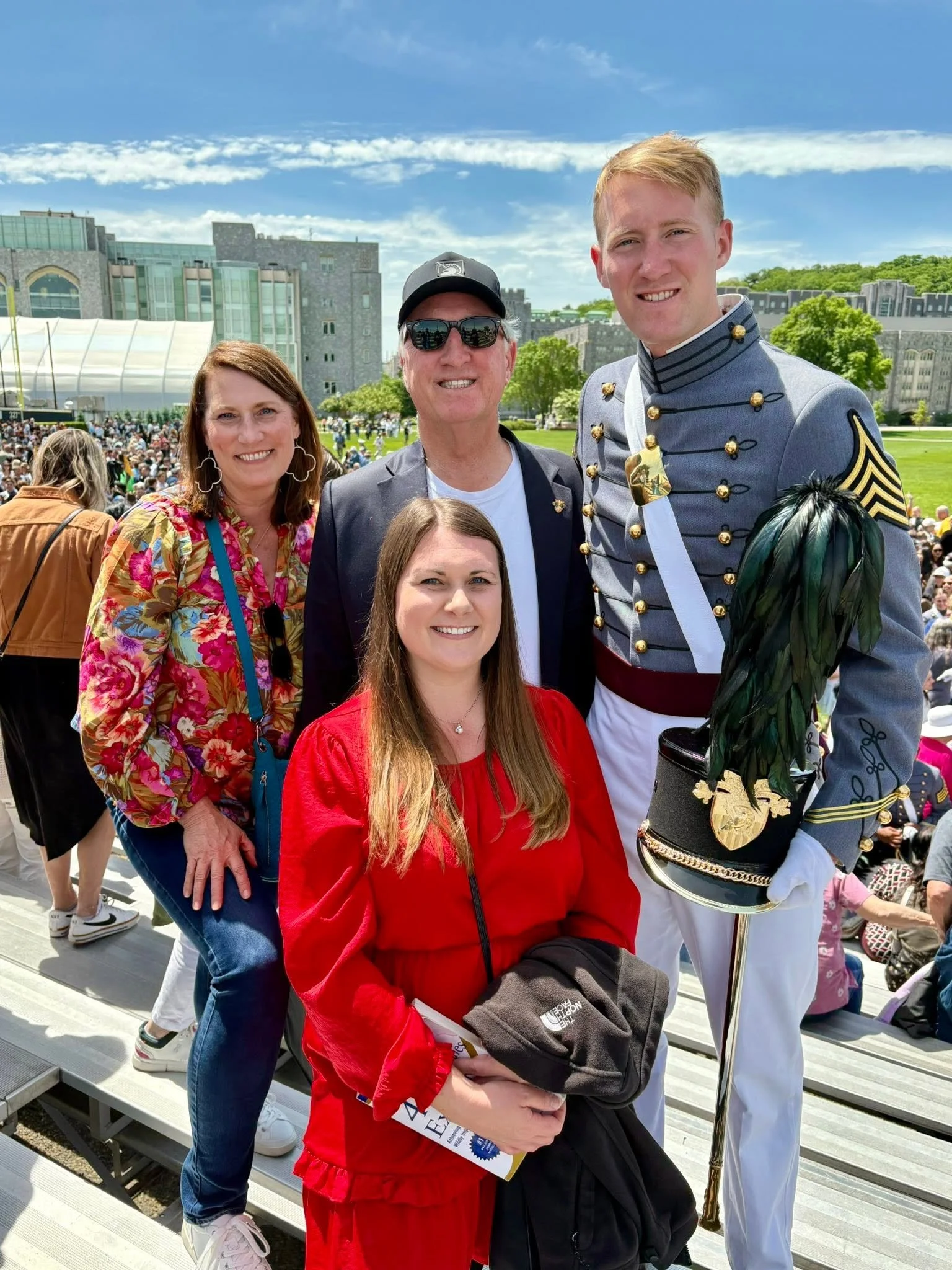 Group of four people smiling at an outdoor event with a large crowd, modern buildings, and green trees in the background. One man is dressed in a marching band uniform, holding a pipe, and the woman in front is wearing a red jacket.