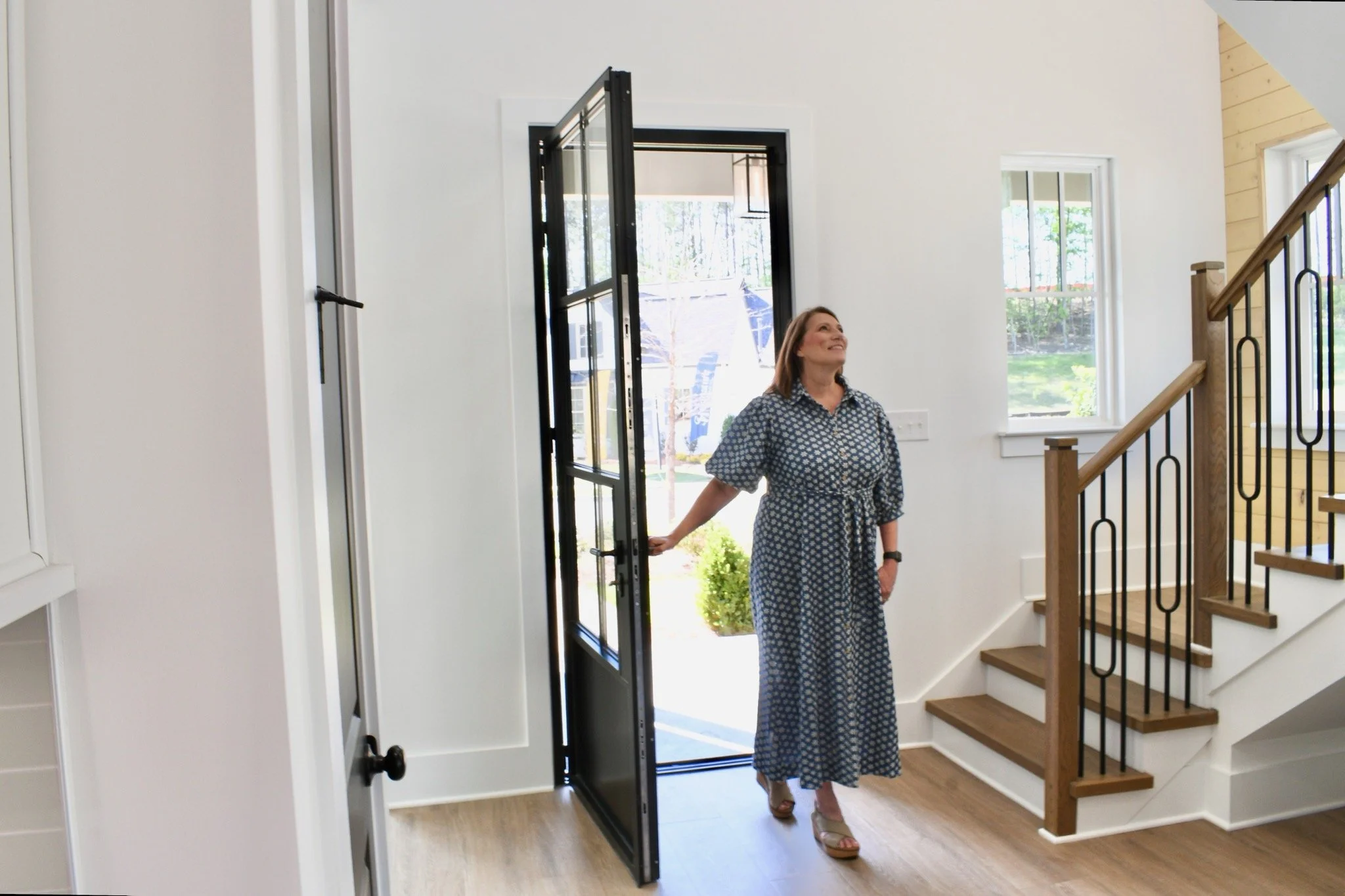 A woman in a patterned dress standing inside a modern home near a glass door, with stairs visible to her right and large windows showing greenery outside.