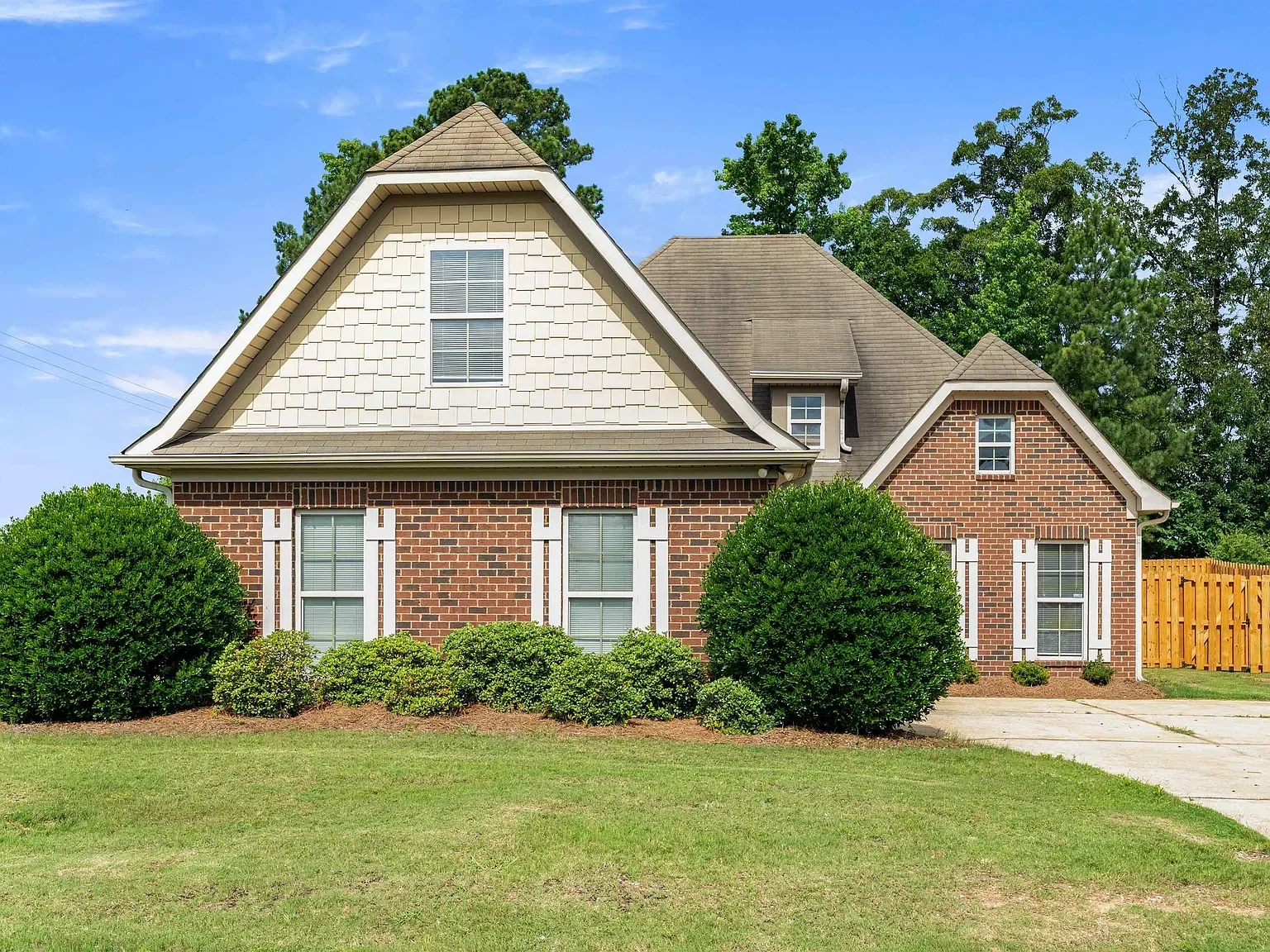 A two-story house with a brick facade and beige siding on the upper level. It has a gable roof, white window shutters, and multiple double-hung windows. The front yard features well-maintained green bushes and grass, with a concrete driveway on the right side.