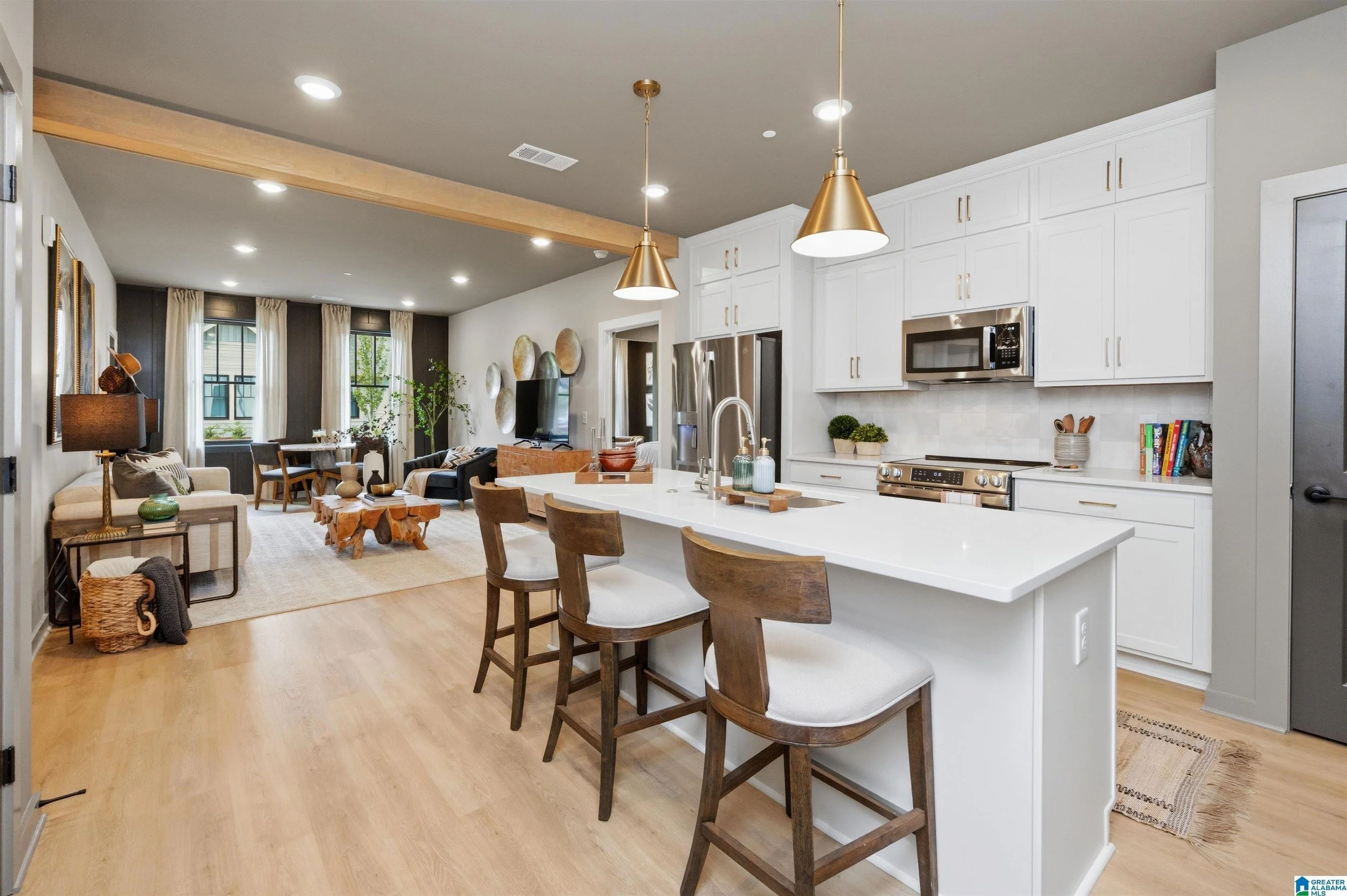 Interior of a modern open-concept living space with a kitchen and dining area, featuring white cabinetry, a large island with bar stools, pendant lighting, hardwood floors, and a sitting area with a sofa, armchair, and decorative wall art.