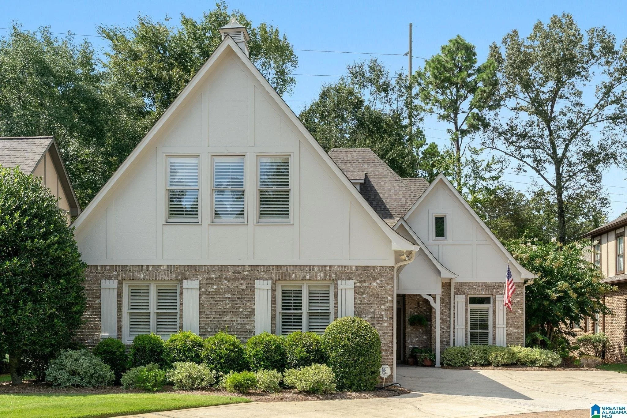 A two-story house with a steep gabled roof, white exterior, and large windows with shutters. The lower part of the house has a brick facade, and there is a small front yard with manicured bushes and a driveway leading to the garage. An American flag is displayed near the entrance, and there are large trees behind the house against a clear blue sky.