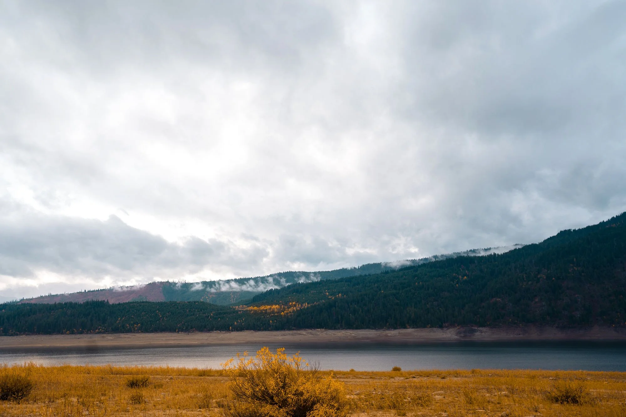 Scenic view of a mountain landscape with Cle Elum Lake in the foreground, forested hills in the middle ground, and cloudy sky overhead.
