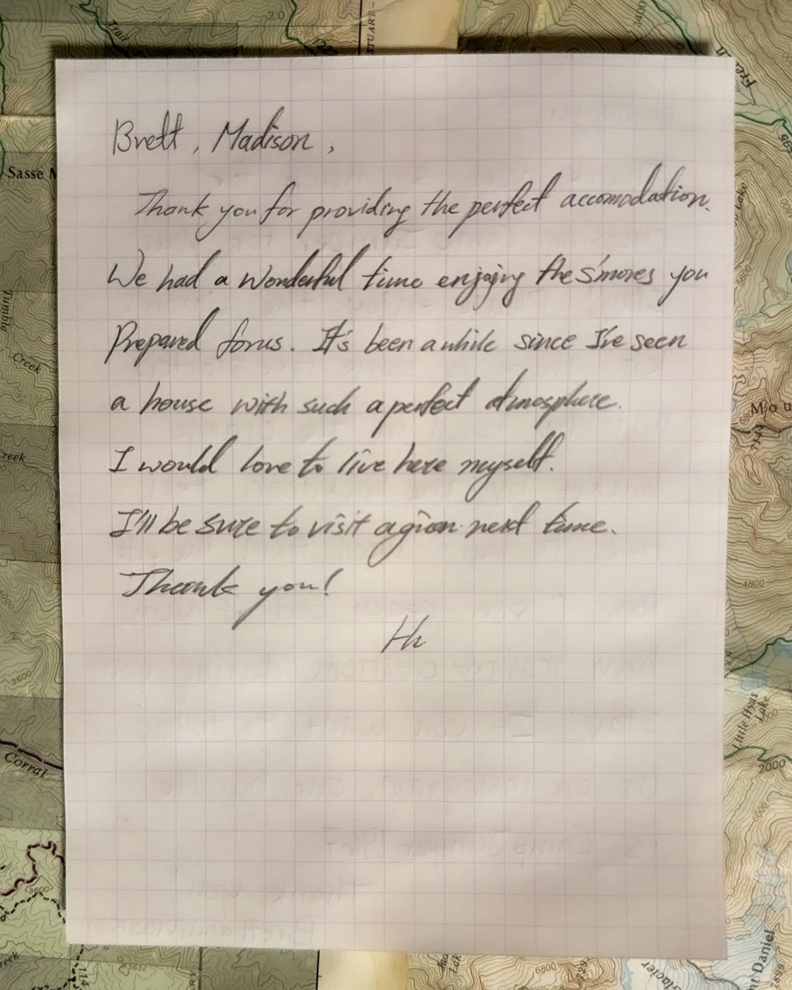 A handwritten thank you note on grid paper, placed on top of a topographical map. The note expresses gratitude to hosts for providing perfect accommodation and mentions enjoying the surroundings, including snow and a house with a good atmosphere.