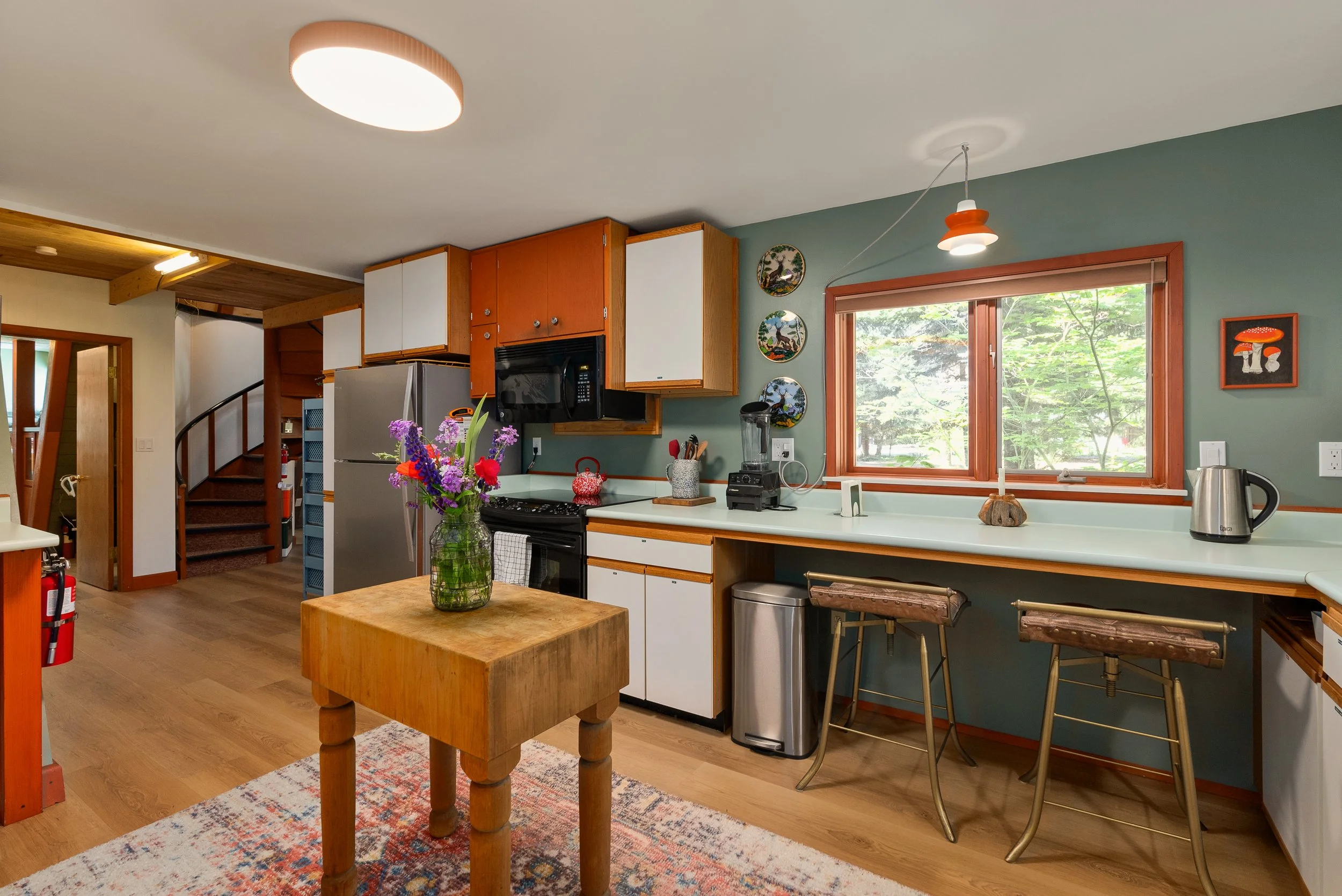 A cozy kitchen with wooden accents, a small wooden table with a colorful flower arrangement, white countertops, modern appliances like a microwave and blender, and decorative wall plates. Large window with views of green trees, and ample natural light with a hanging lamp and a ceiling light.