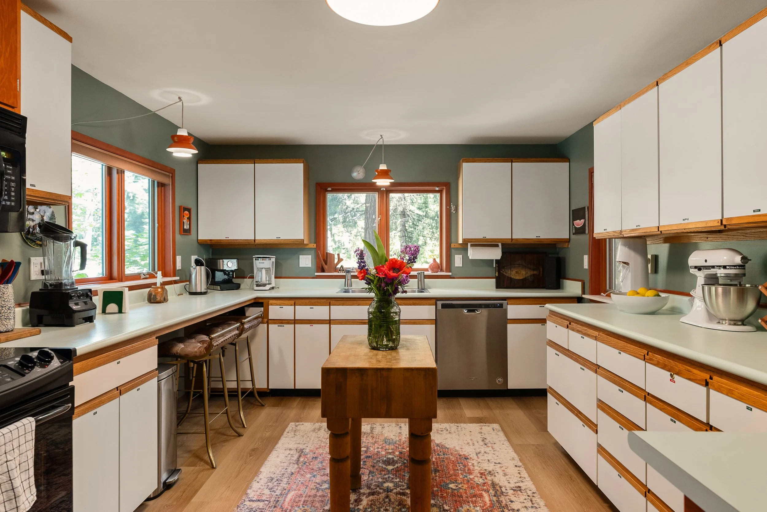 A spacious kitchen with white cabinets and wooden trim, large windows overlooking greenery, a central wooden table with a glass vase of colorful flowers, and various appliances including a coffee maker and mixer.
