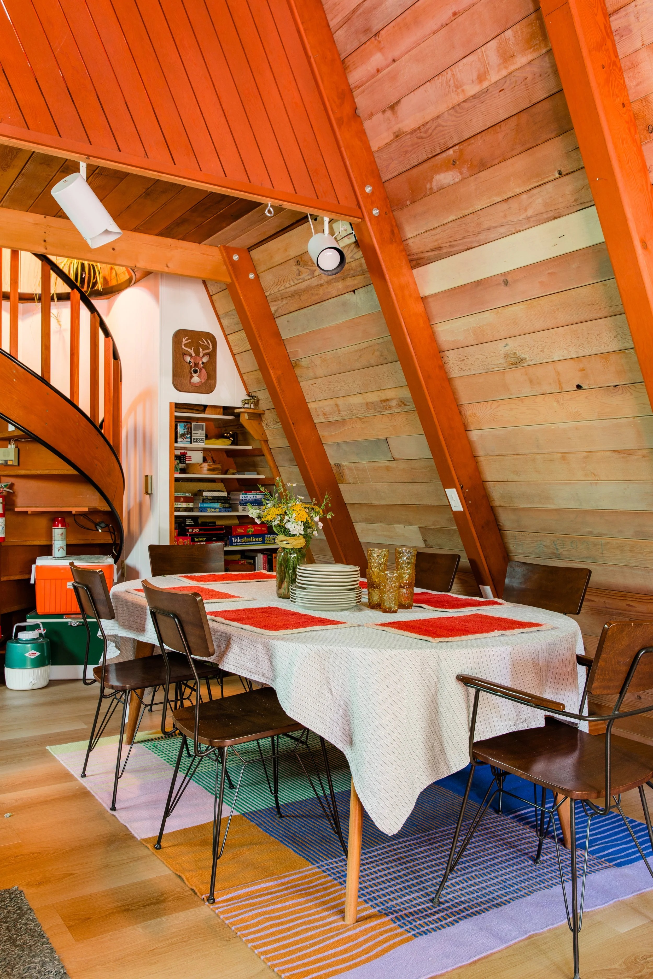 A-Frame cabin dining room with a wooden sloped ceiling, a white tablecloth on the dining table, a vase with flowers, stacked plates, and vintage glasses. Wooden chairs surround the table, and a colorful striped rug is beneath it.
