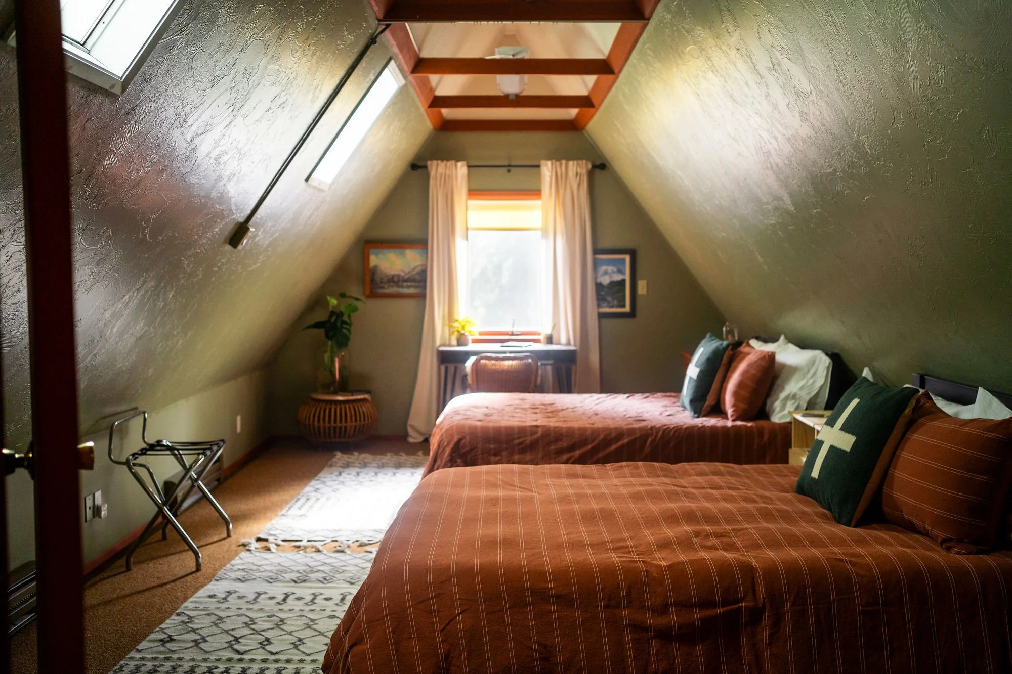 A bedroom with two beds covered in orange and brown checkered bedding, a window with white curtains, framed paintings on the wall, a small desk with a chair, a potted plant, and a rug on the floor.