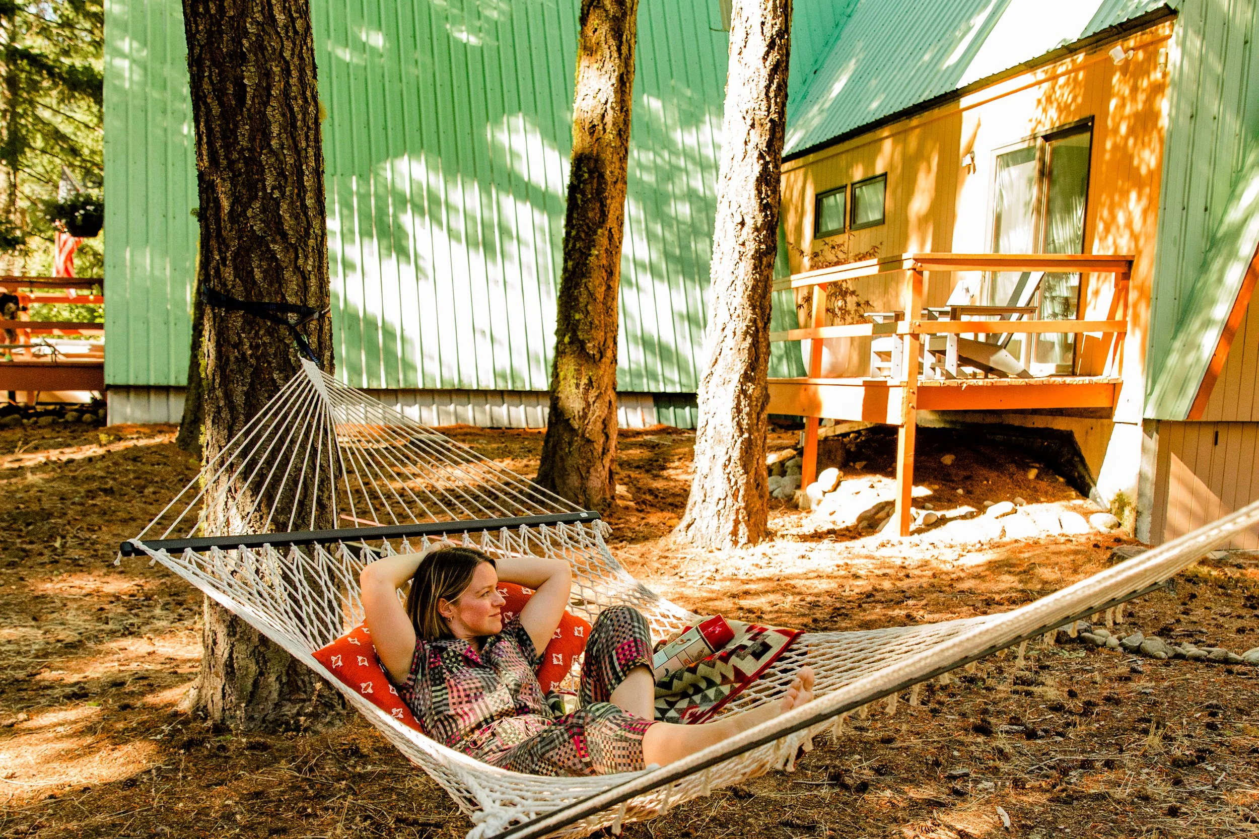 A woman relaxing and reading a book on a hammock tied between trees in a wooded area beside a colorful building with green and yellow siding and a small wooden deck with adirondeck chair.