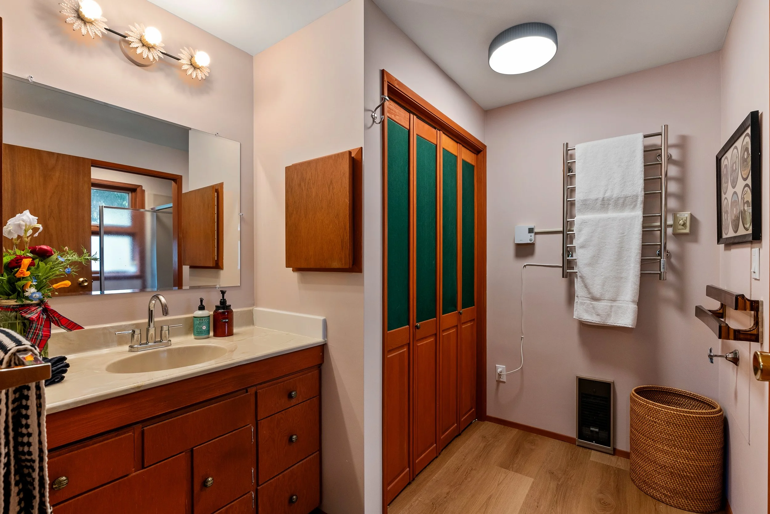 A bathroom with a wooden vanity, a mirror, a sink, flower decorations, soap and lotion bottles, a wooden cabinet with green panels, a towel rack with a white towel, a framed picture, and a wicker laundry basket.