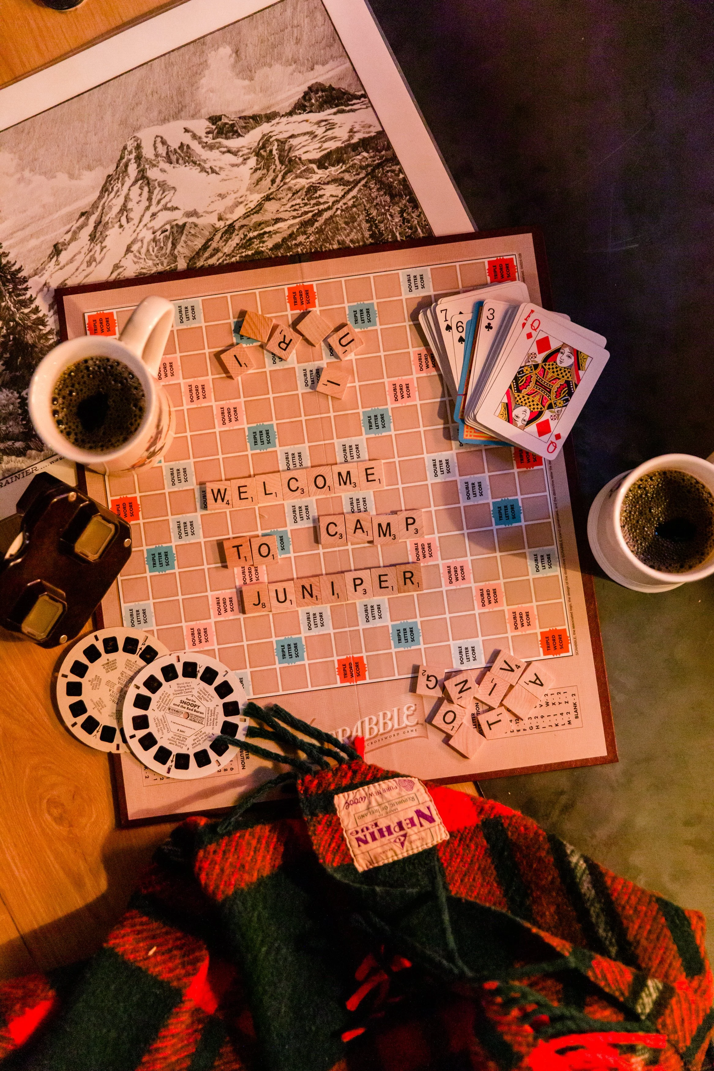 A tabletop with a scrabble board and game pieces, two cups of coffee, and playing cards. The Scrabble letters spell out 'WELCOME TO CAMP JUNIPER.' There is a mountain illustration in the background view.