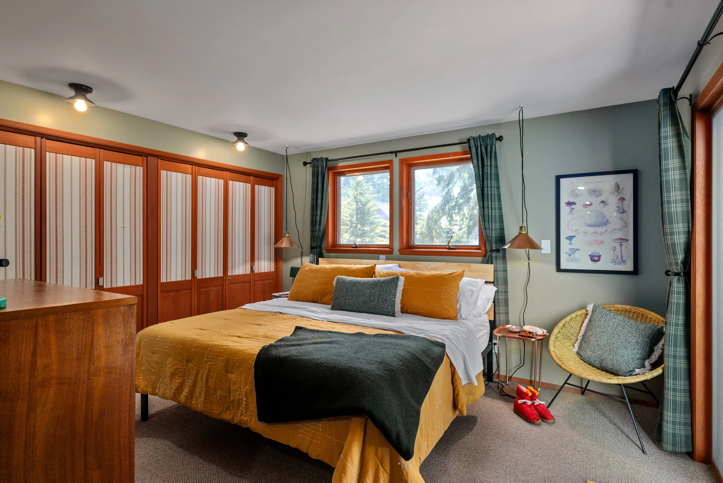 Photo of a cozy bedroom with a large bed covered in mustard yellow bedding and green wool blanket, multiple pillows, and a window with green plaid curtains.