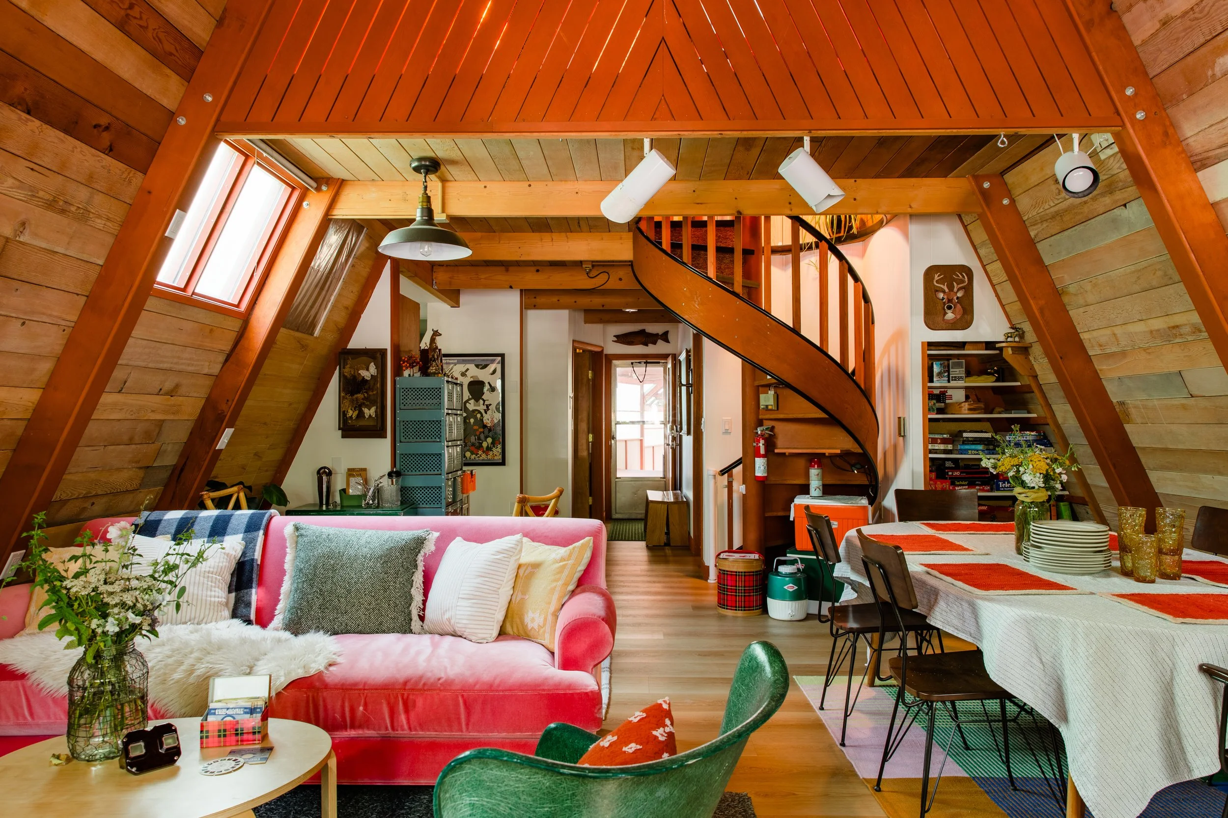 Interior of a cozy wooden home with a loft, featuring a pink sofa with colorful pillows, a round coffee table with magazines and decorative items, a dining table with plates and glasses, and a staircase leading to a upper level. The room has wood-paneled walls and ceilings, large windows, and decorative animal artwork on the walls.