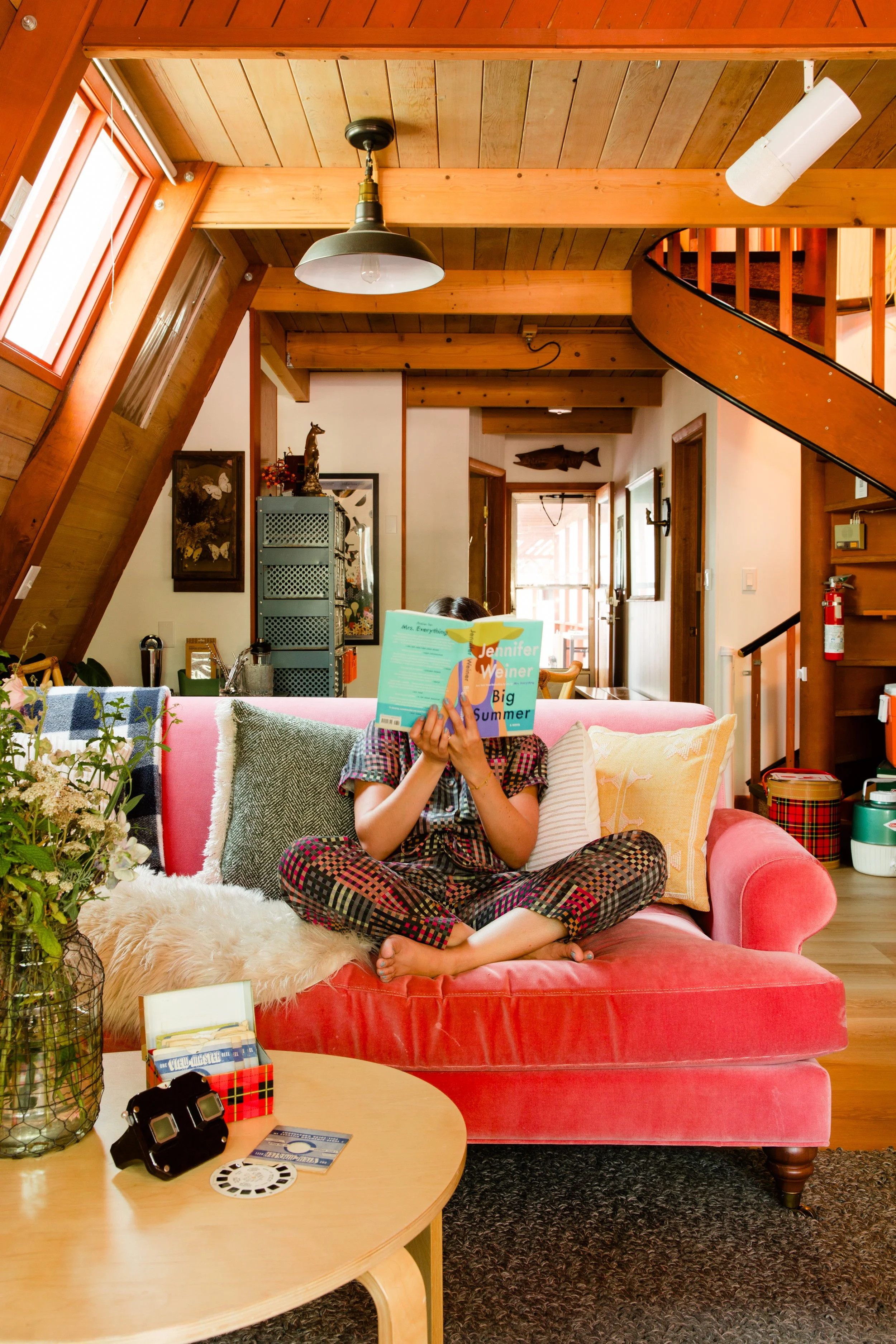 A person sitting cross-legged on a pink velvet couch, reading a book, in a cozy, wood-paneled A-Frame cabin living room.