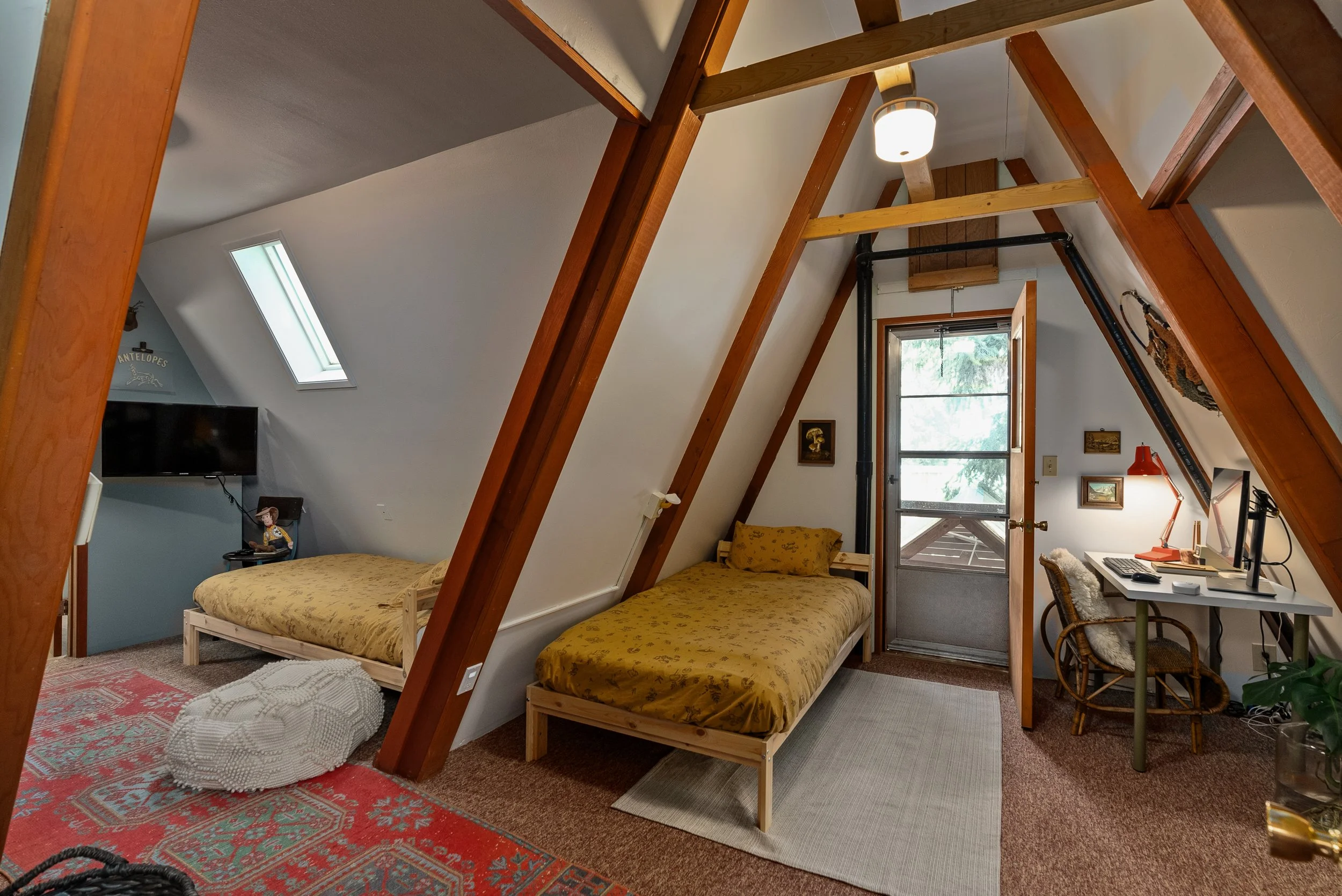 Upstairs room with two twin beds with yellow bedding, wooden beams, a desk with a computer and a wicker chair, a window, and a skylight, decorated with pictures and a lamp, carpeted floor.