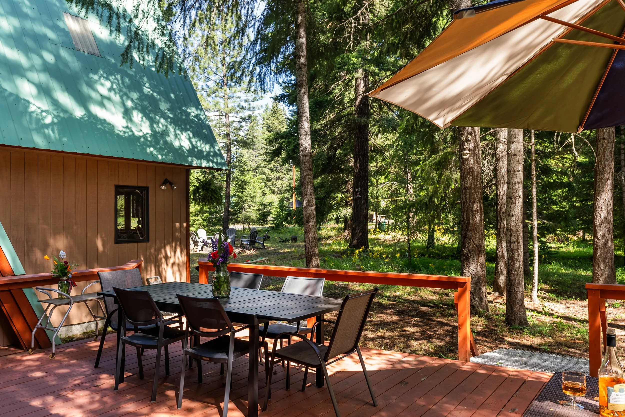 Outdoor wooden deck surrounded by trees with a patio table, chairs, and an umbrella, next to a cabin with a teal metal roof and a window, amidst a forest setting.