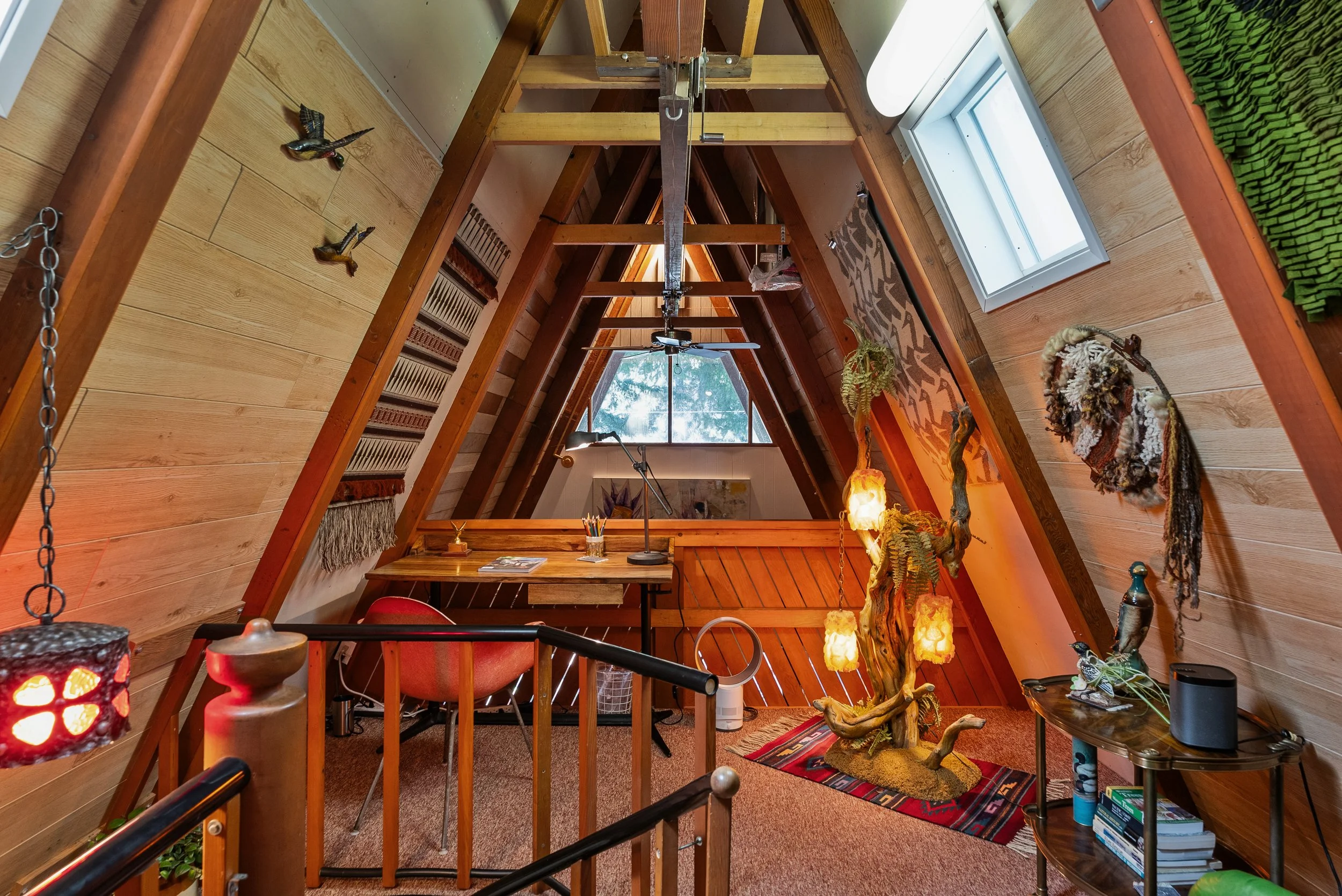 Interior view of an A-frame loft with wood-paneled walls, a skylight window, and various decorative items including hanging sculptures and sculptural vintage driftwood lamp.