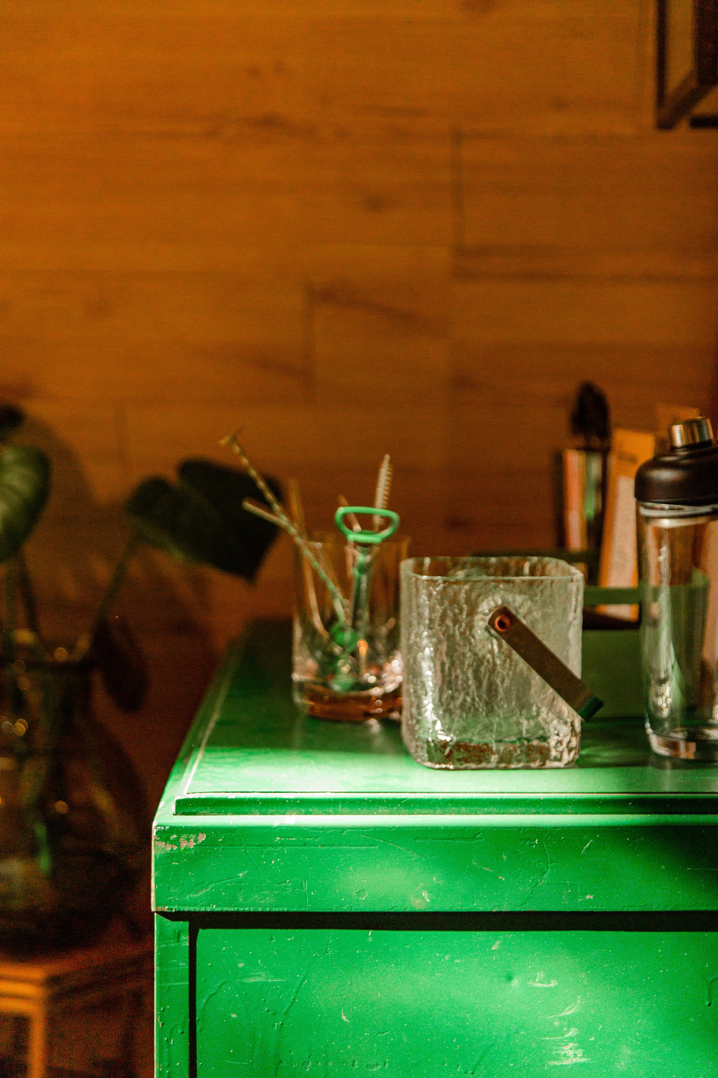 A green shelf with ice bucket and cocktail mixing containers on it. The background is brown wood with some objects including a book and a bottle.