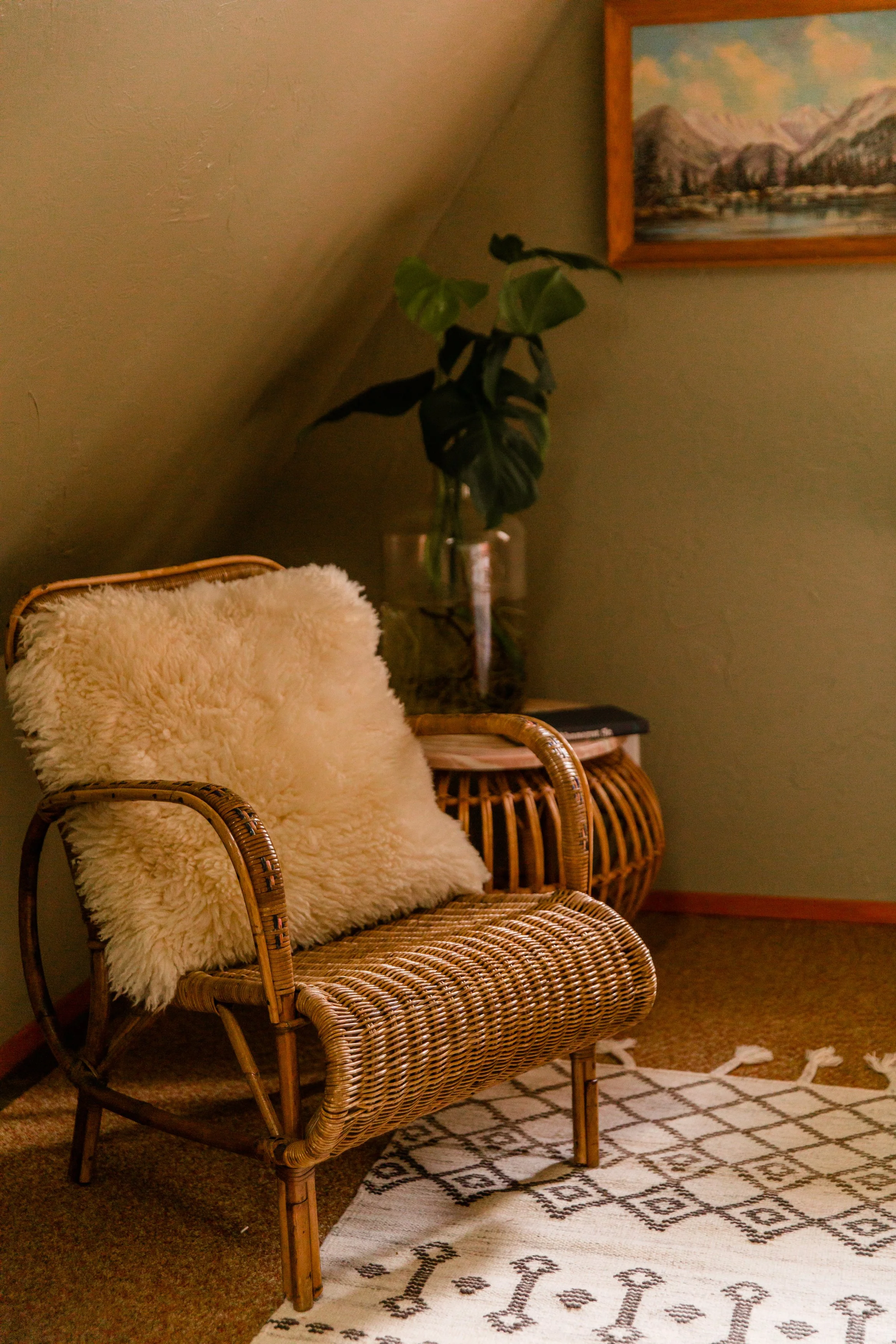 A French wicker chair with a sheepskin pillow. There is a vintage bamboo table with a plant behind the chair, and a framed landscape painting with mountains and a lake on the wall. The room has a patterned rug, and green-painted walls.