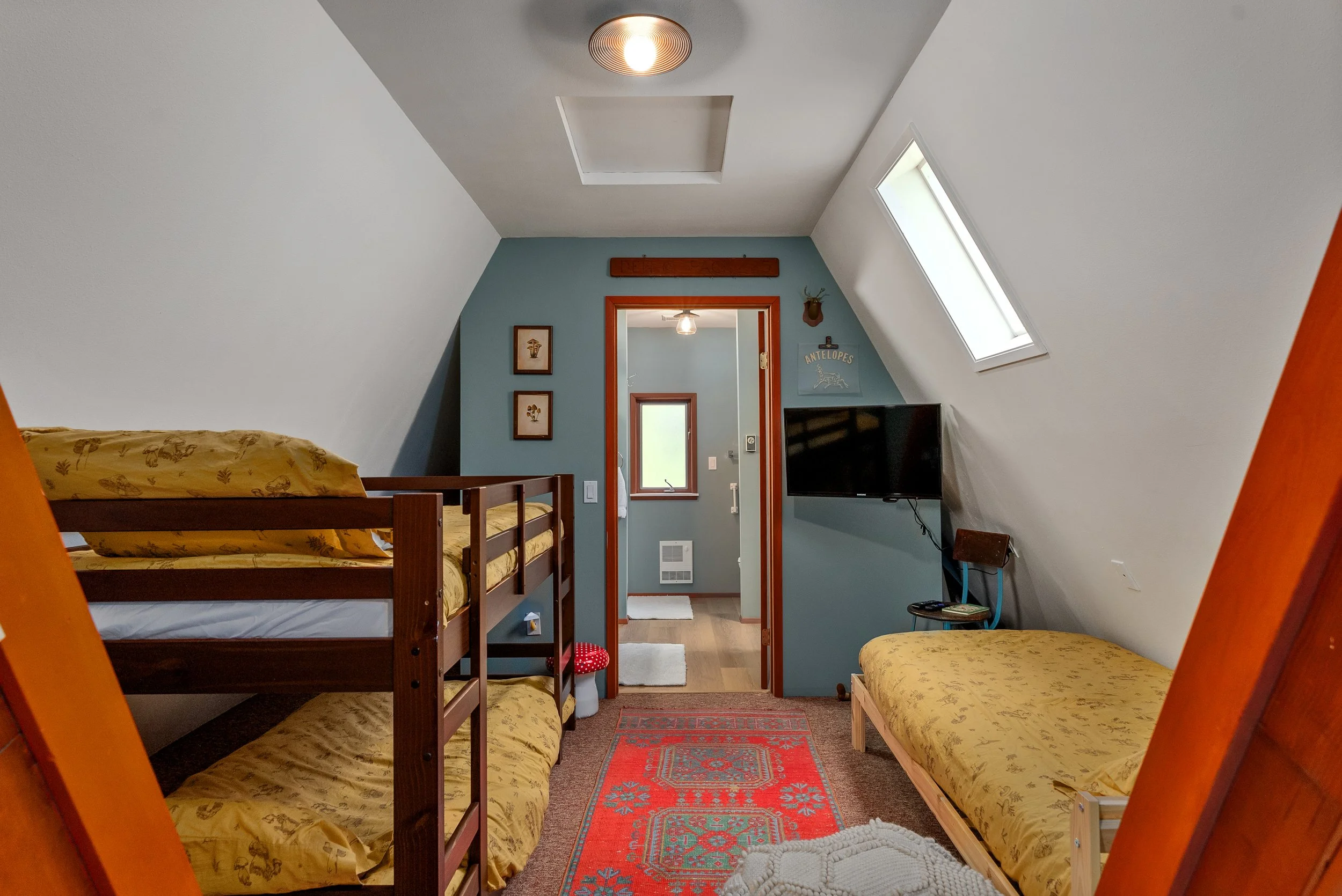 A cozy upstairs bedroom with two beds with yellow floral bedding, a flat-screen TV on the wall, framed artwork, a red patterned rug, and a skylight window in the sloped ceiling.