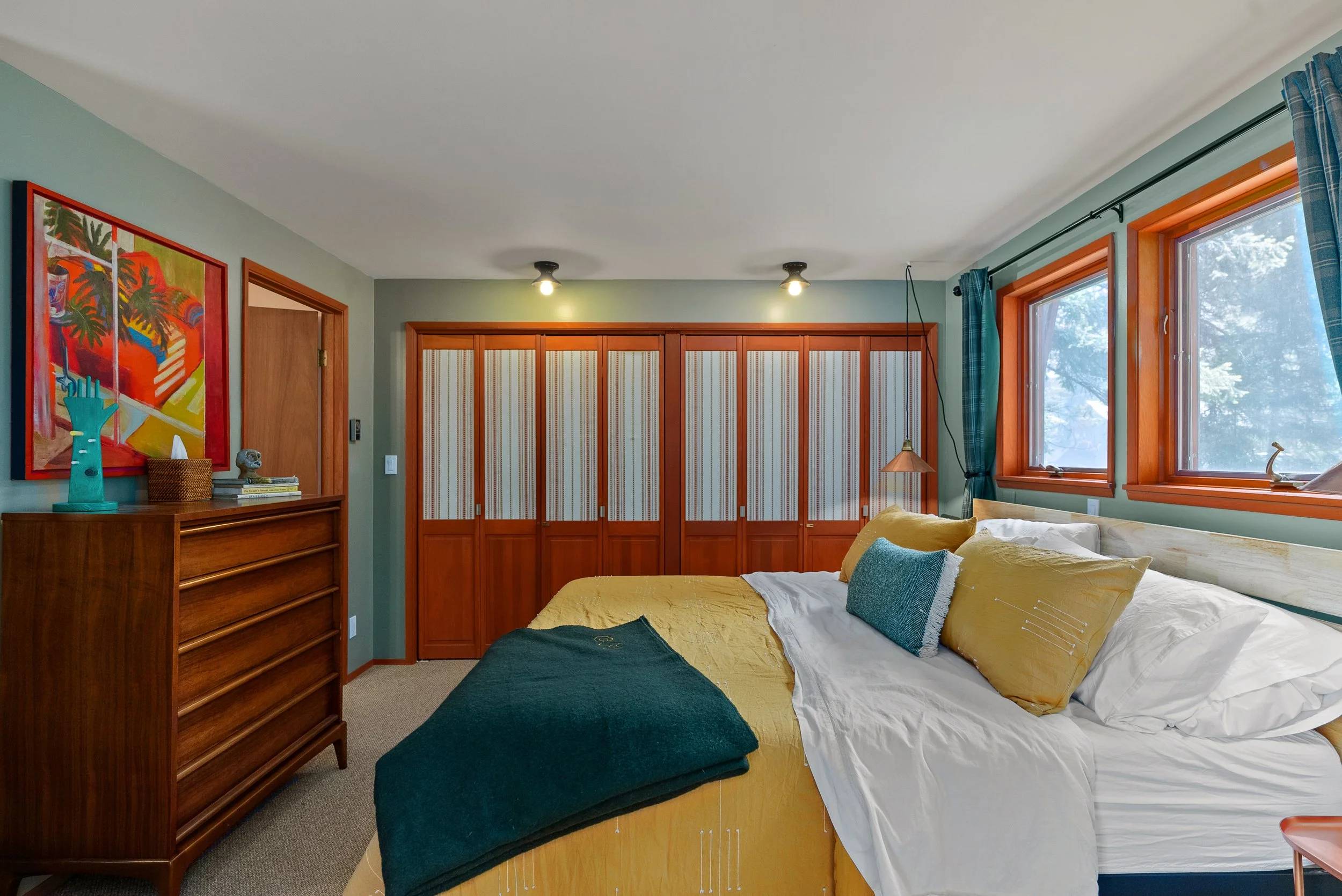 A cozy bedroom with a large bed featuring yellow and white bedding and decorative pillows, vintage wooden furniture, colorful artwork on the wall, a window with blue curtains, and a built-in wardrobe with striped doors.